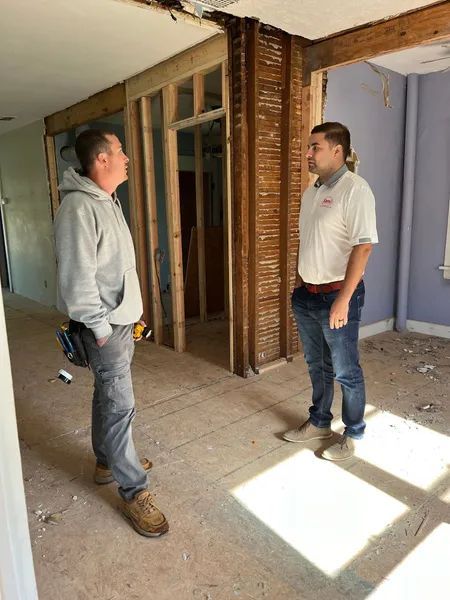 Two men in a room under renovation, discussing construction plans. One in a gray hoodie, the other in a white shirt.