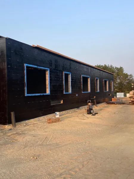 Black building exterior with open window frames; a person works nearby on a gravel lot.