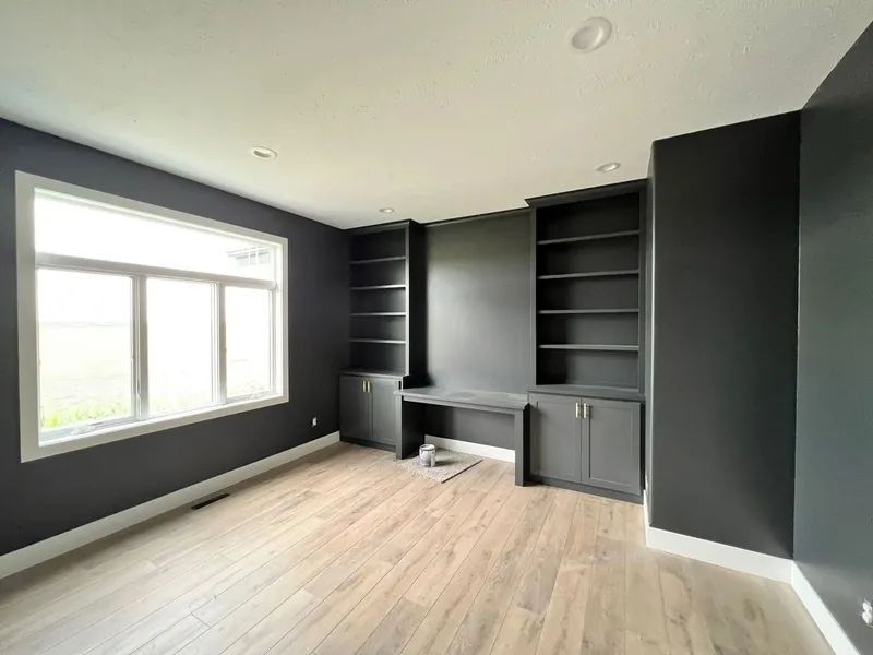 Empty home office with dark gray walls, built-in desk, and window. Light wood floor and white trim.