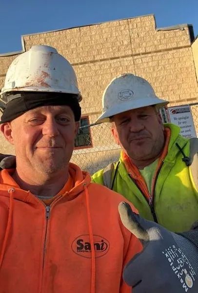 Two construction workers in hard hats and safety vests smiling; one gives a thumbs up.