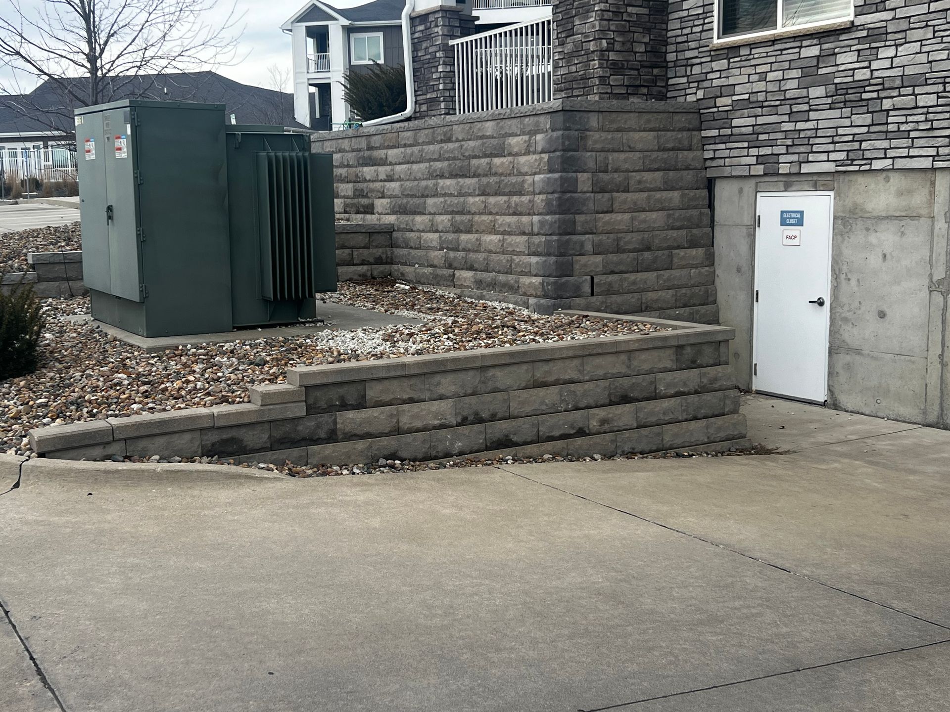 Outdoor scene with electrical equipment and a retaining wall near a building entrance and parking area.