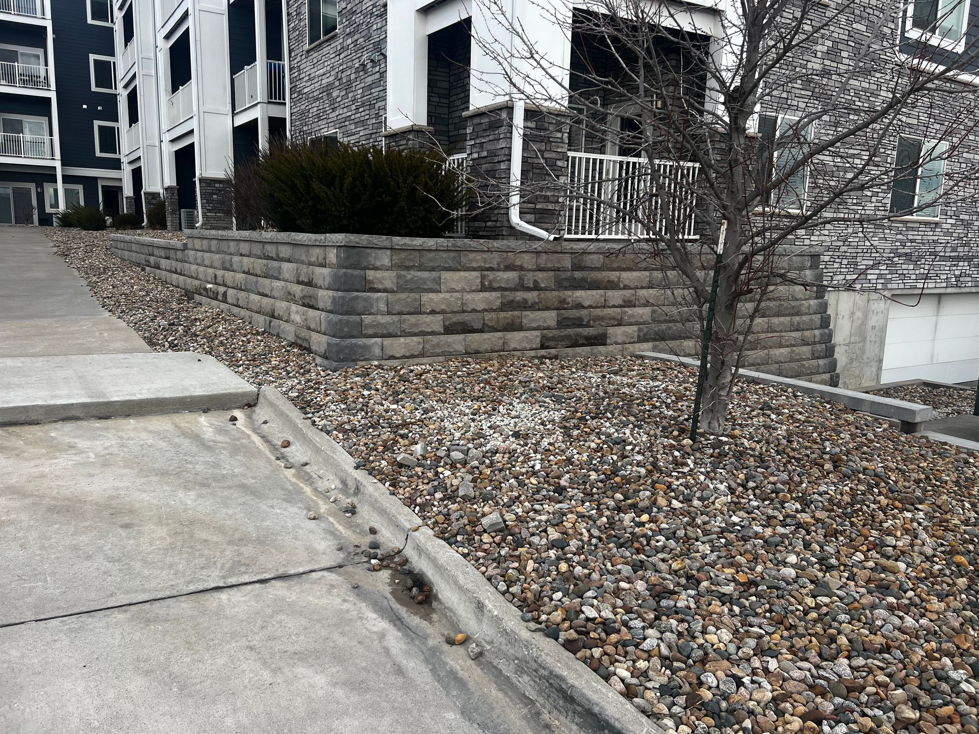 A retaining wall made of gray blocks next to a building with pebble ground cover. A sidewalk is in front.