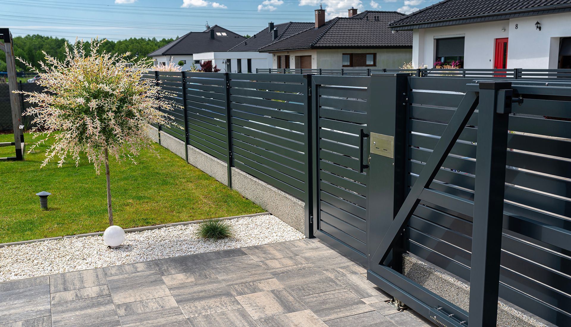 Dark gray slatted fence with gate and small tree in front of several houses.