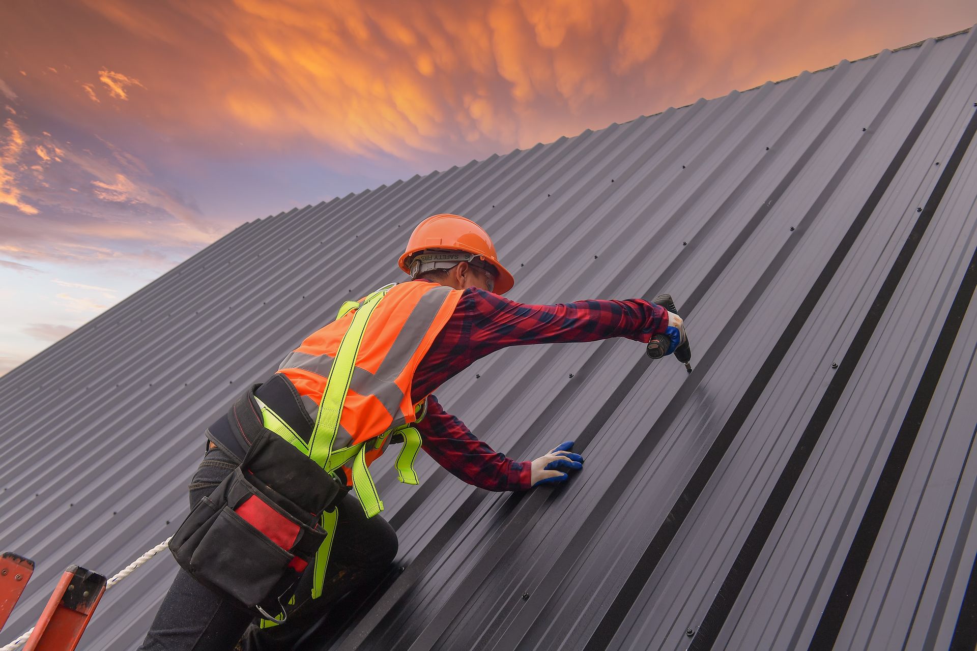 Worker in safety gear installing a metal roof at sunset on a sloped building.