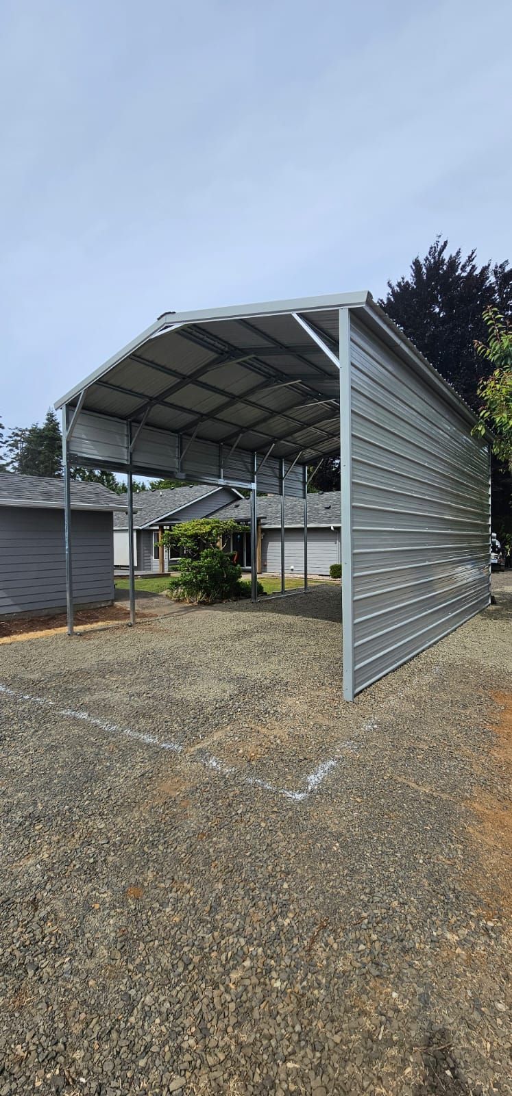 A large gray metal carport in a gravel area, with some houses and trees in the background.