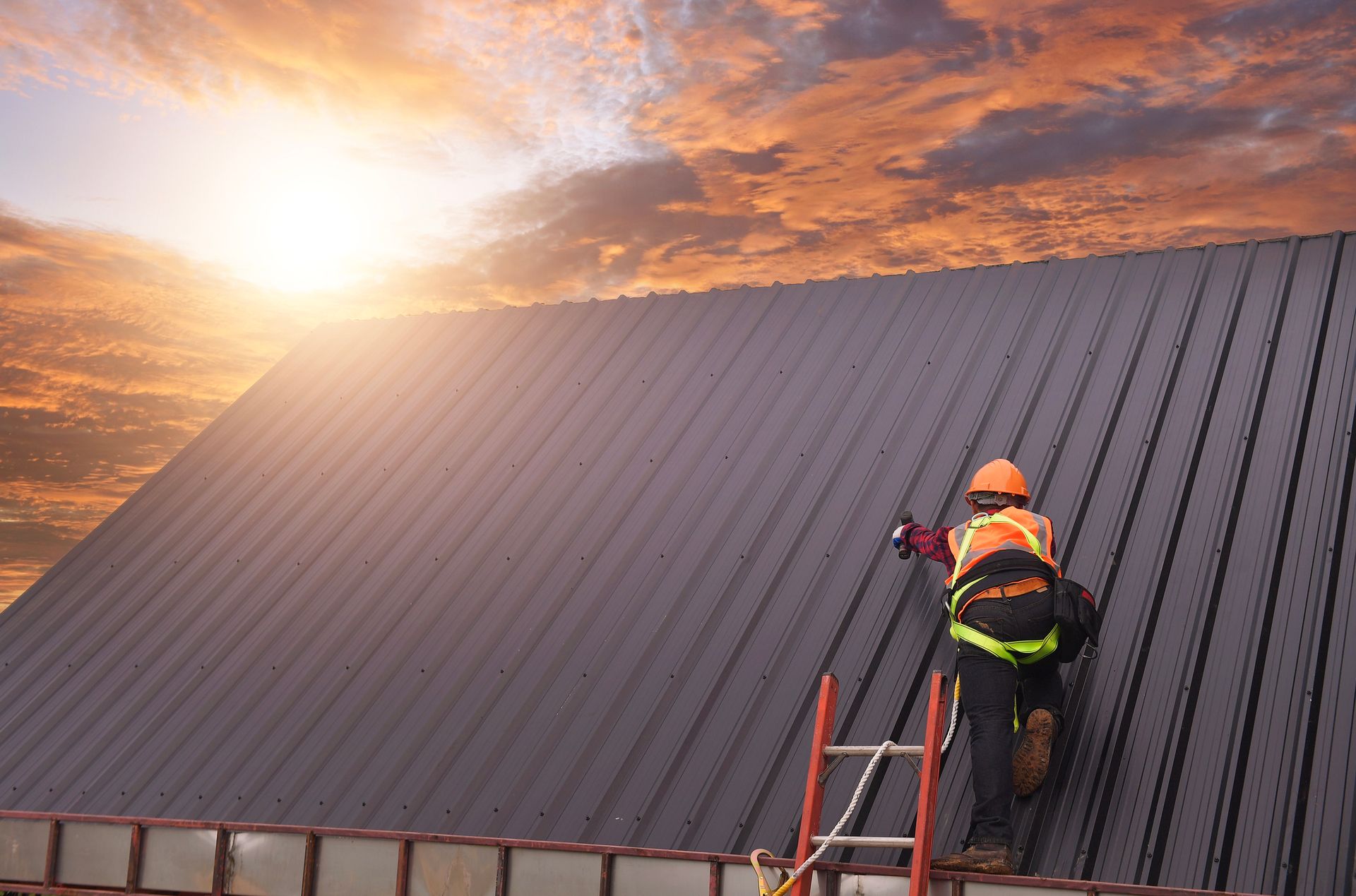 Worker in safety gear inspects metal roof on ladder at sunset.