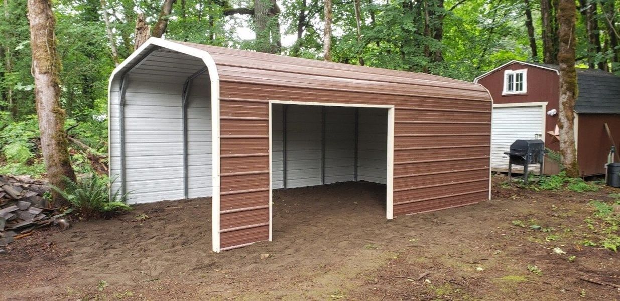 A brown and white metal garage with a barn in the background.