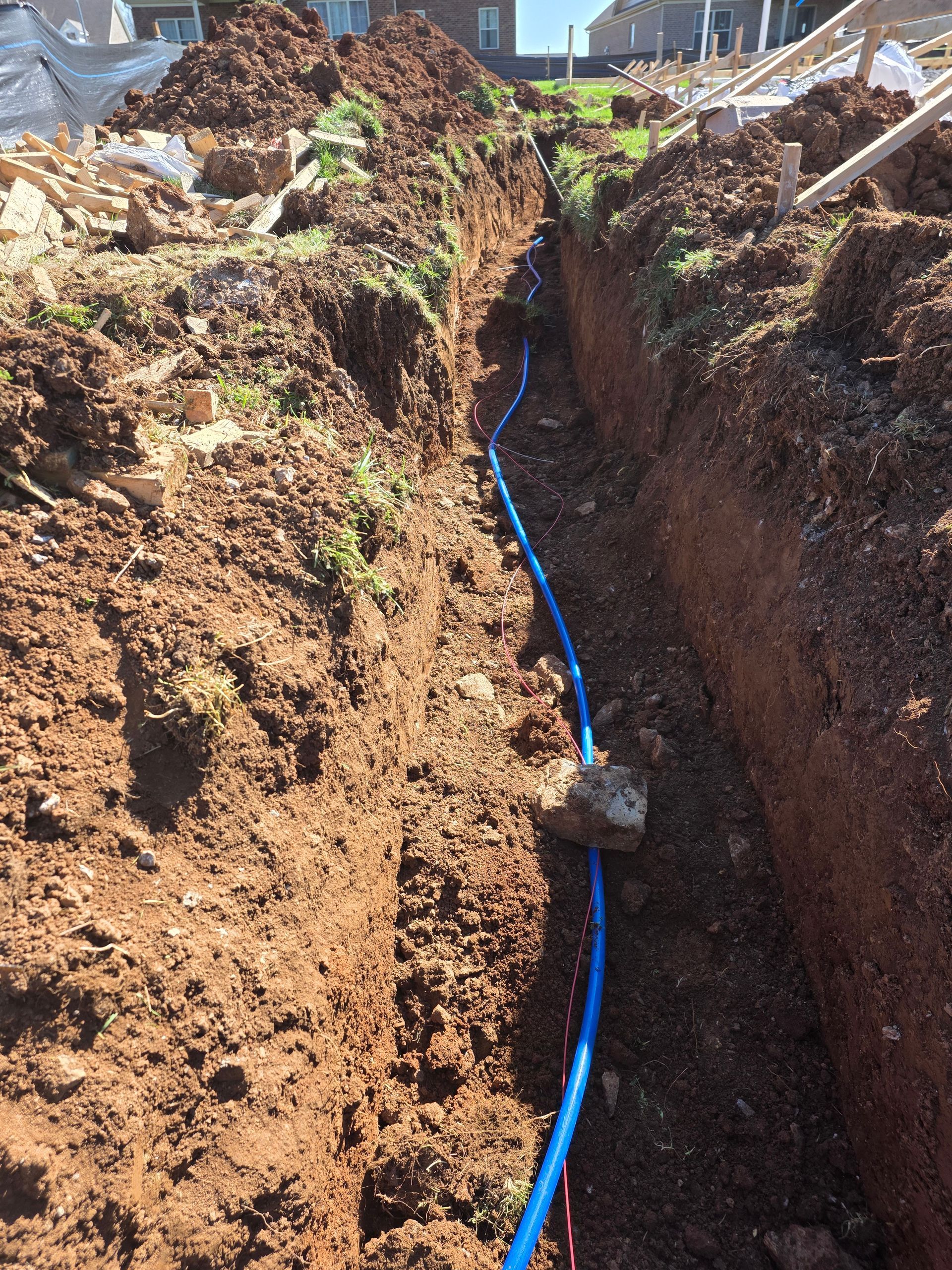 Deep trench in reddish soil with a blue pipe laid along the bottom at a construction site