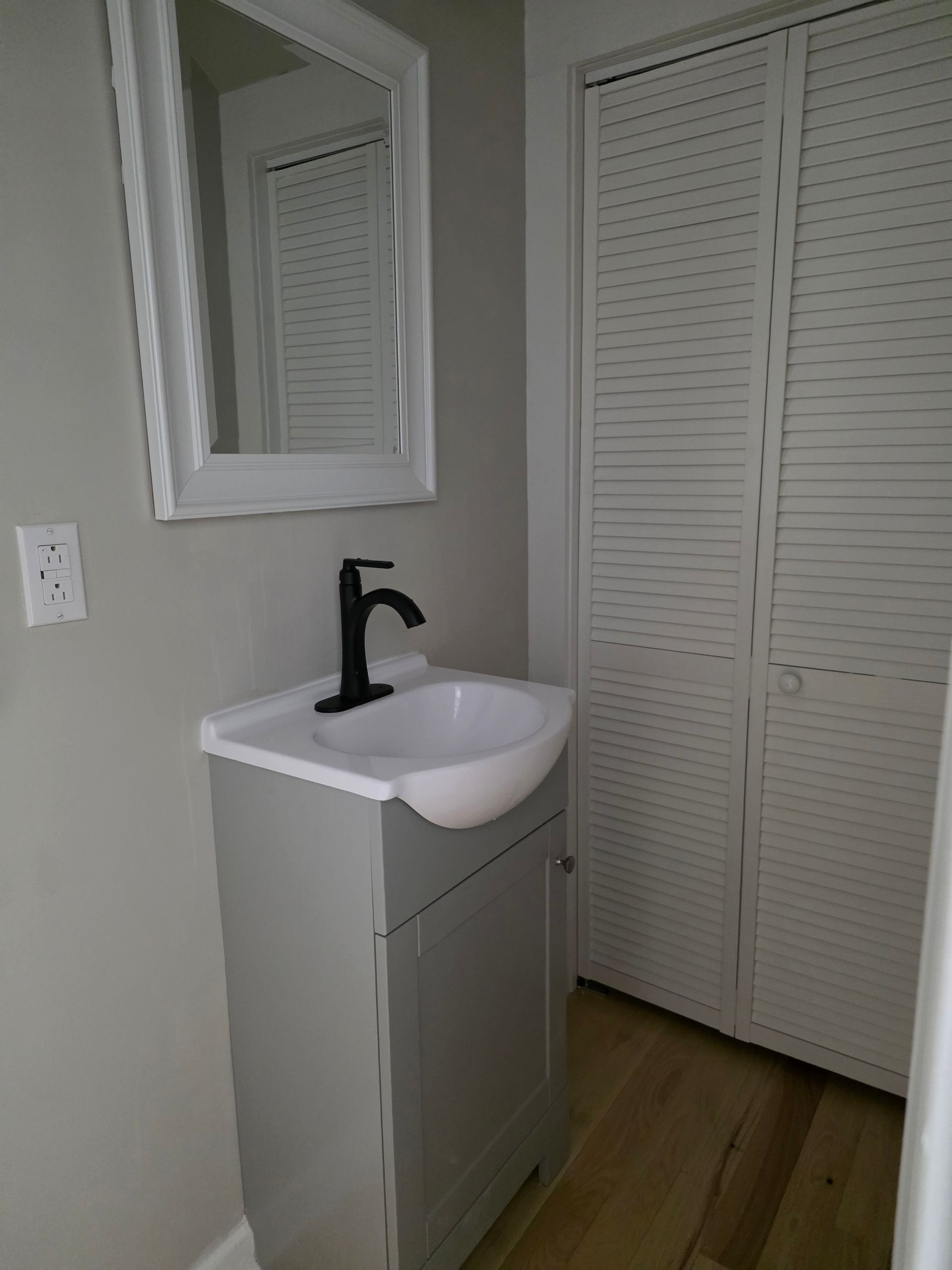 Small bathroom vanity with white sink, black faucet, mirror, and louvered closet doors.