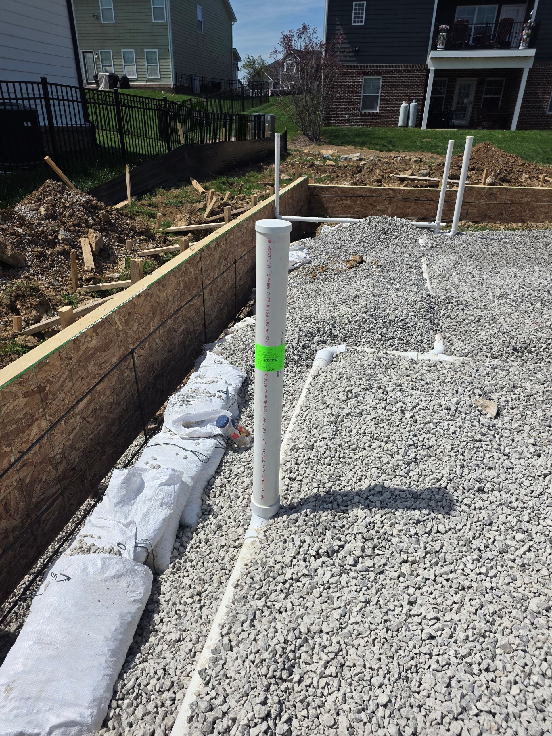 White PVC pipe marker beside a gravel foundation trench with cinder blocks and wooden edging in a yard