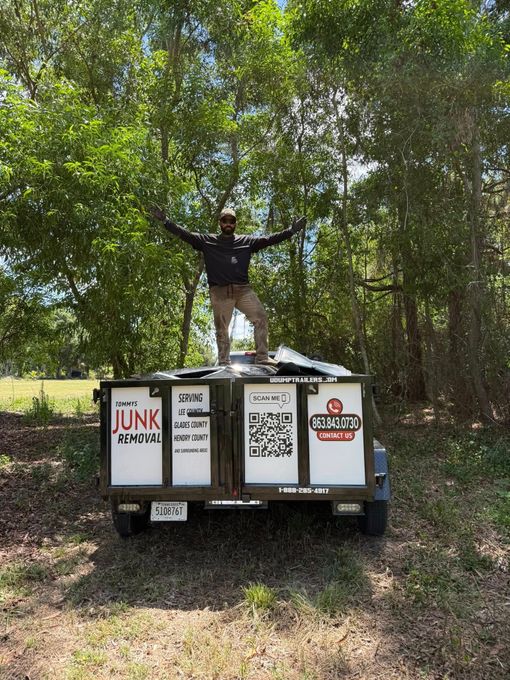 Man standing on junk-filled trailer with arms raised in a wooded area. Signs on trailer read