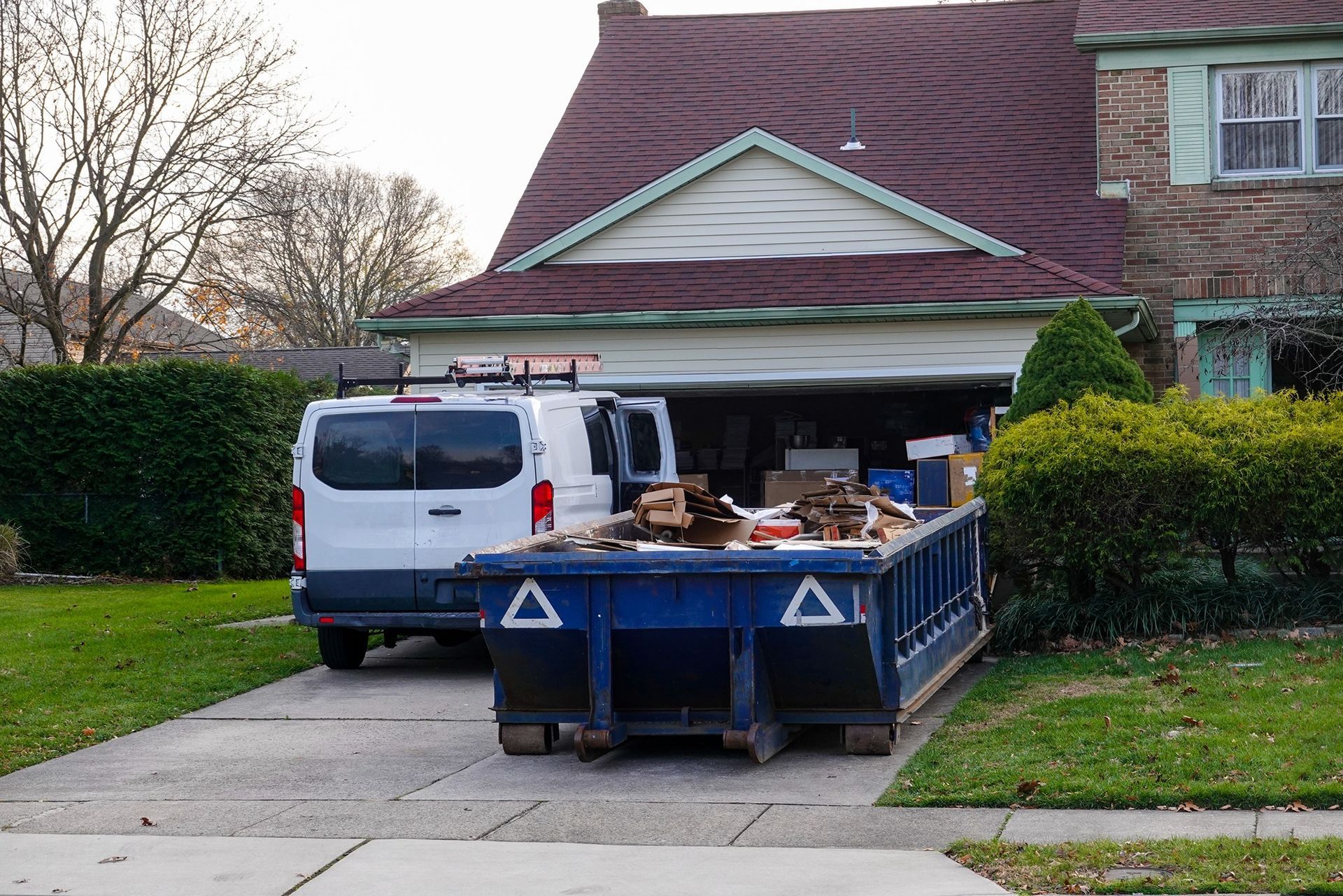 A blue dumpster filled with debris sits in a driveway next to a white van and a house with an open garage.