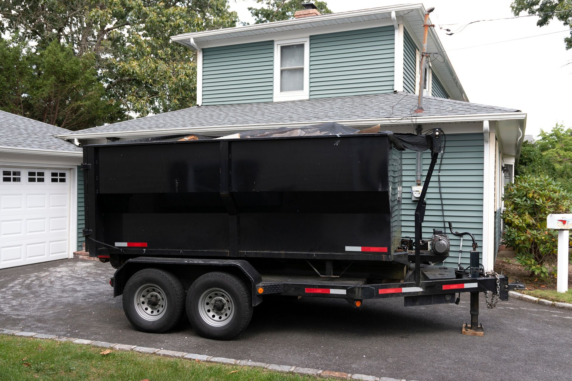 Black dumpster trailer parked in front of a blue house and white garage.