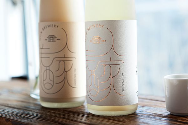 나무 테이블 위에 놓인 두 개의 사케병. 하나는 옅은 베이지색이고 다른 하나는 흰색이며, 우아한 금색 라벨과 작은 흰색 컵이 달려 있습니다.