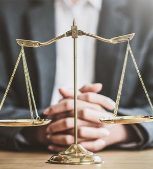 A man is sitting at a table with his hands folded in front of a scale of justice.