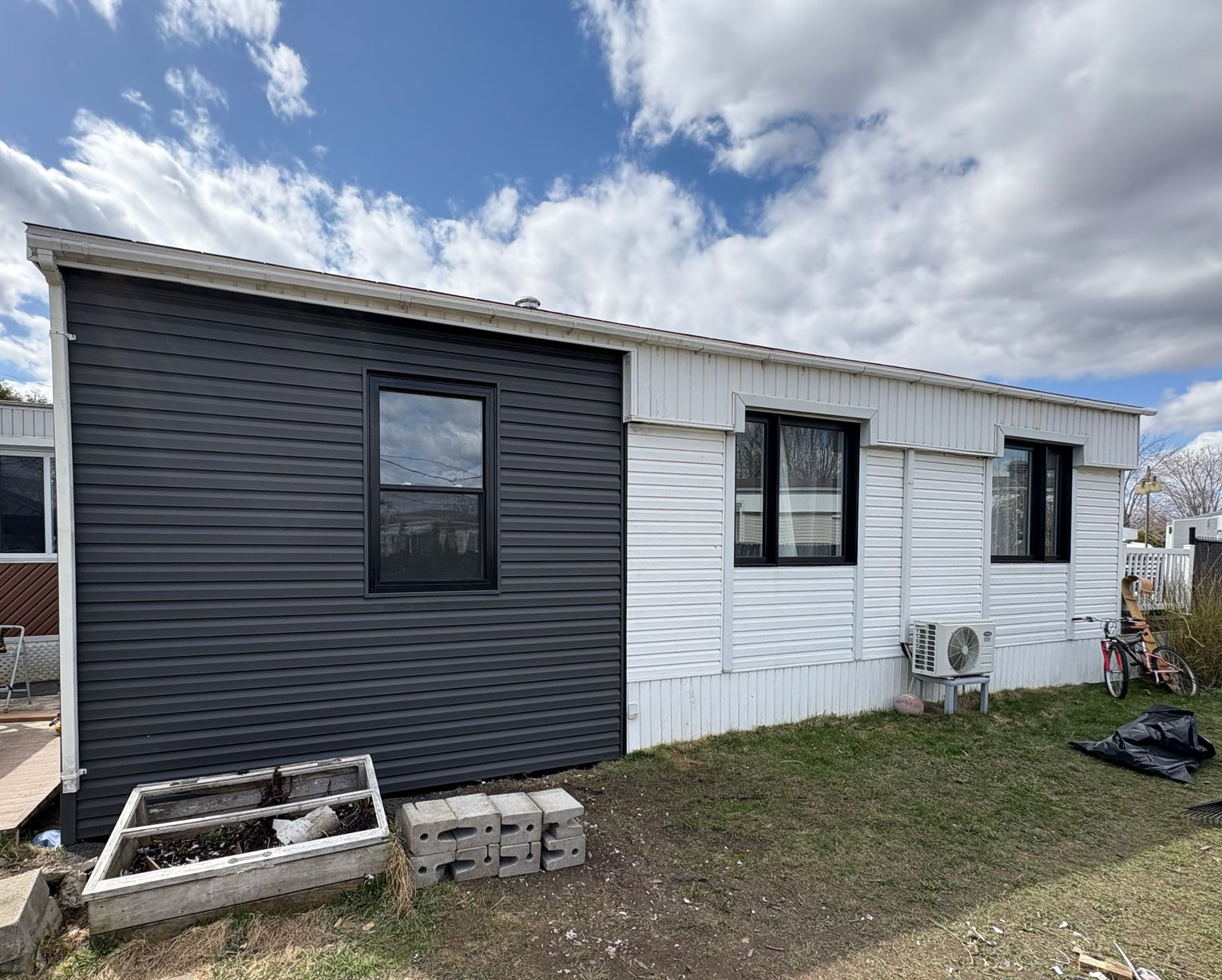 Extérieur d'une maison avec un revêtement noir d'un côté et blanc de l'autre, fenêtres et unité de climatisation.