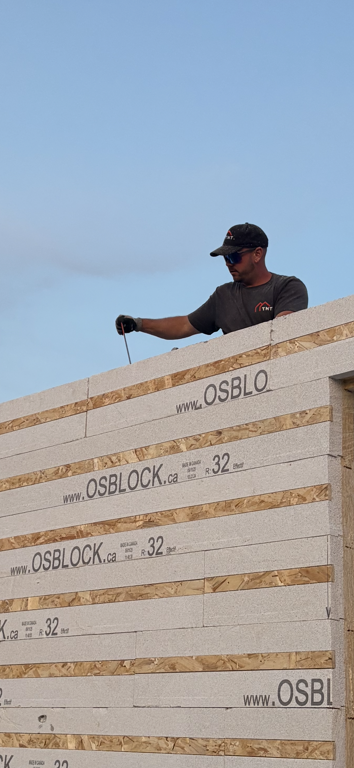 Un ouvrier du bâtiment, sur un toit, mesure le revêtement en panneaux OSB à l'aide d'un mètre ruban. Ciel bleu en arrière-plan.