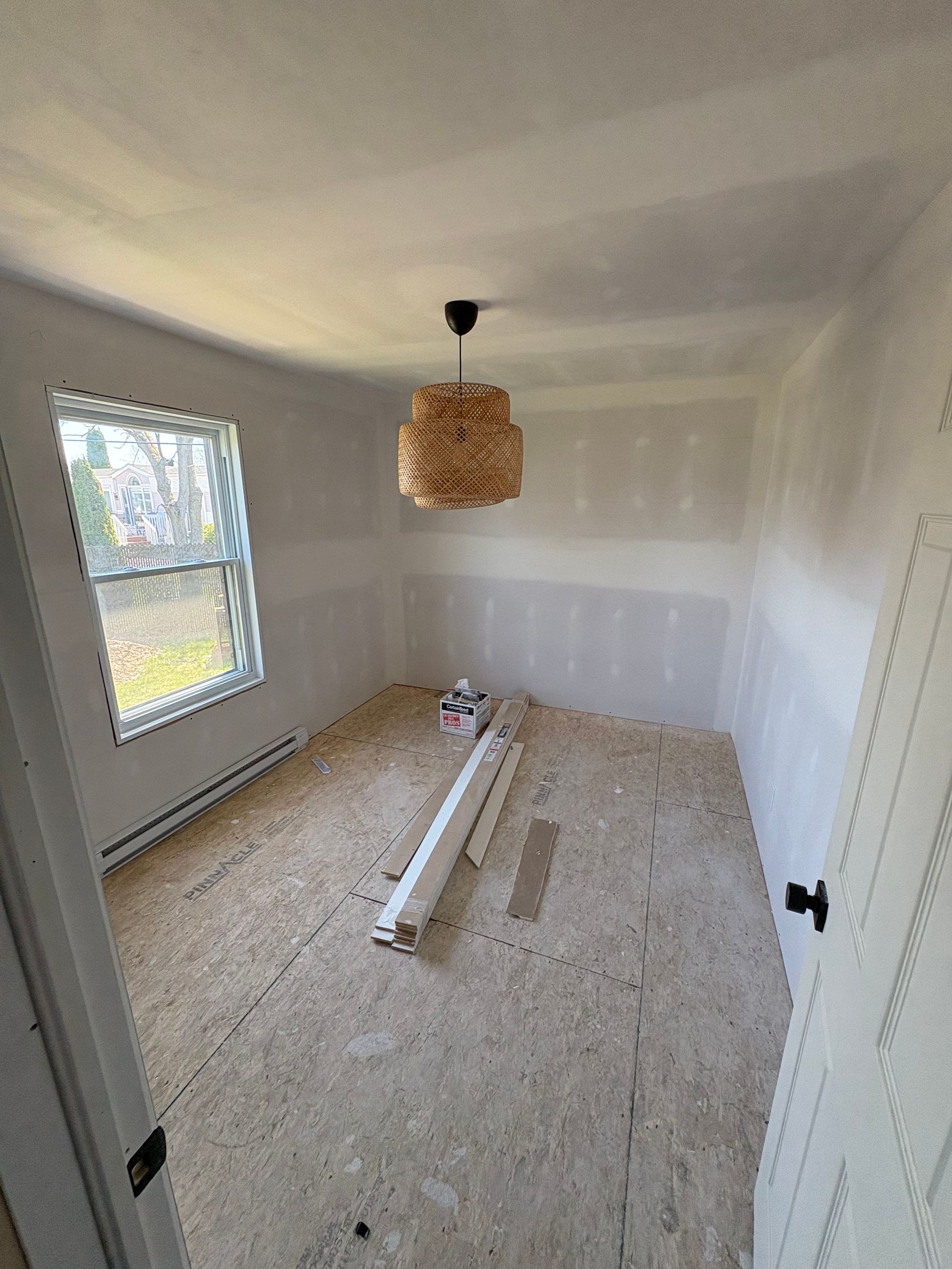 Pièce inachevée avec cloisons sèches, fenêtre, parquet nu et luminaire décoratif.