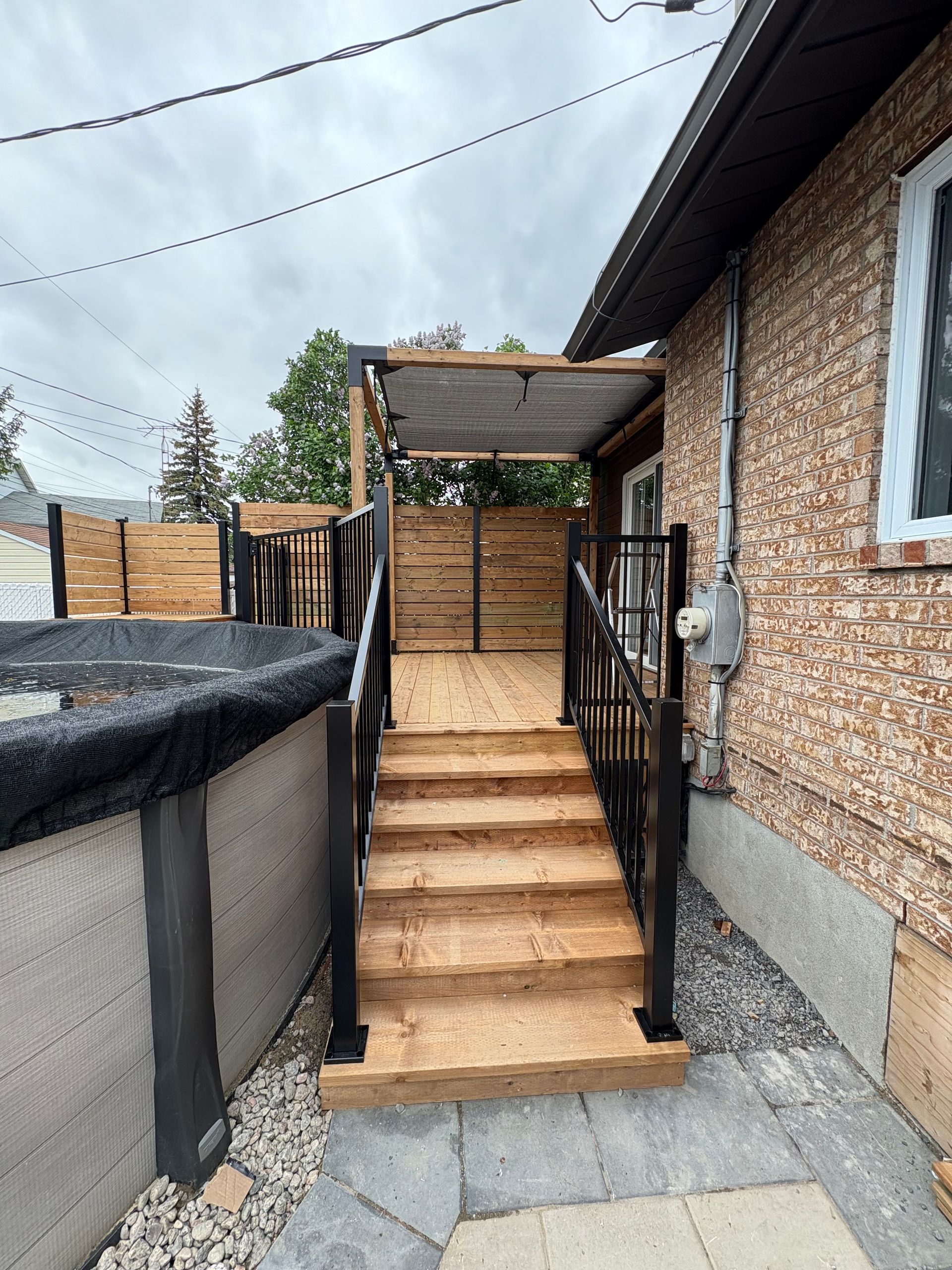 Terrasse en bois avec escalier menant à un espace ombragé, à côté d'un bâtiment en briques et d'une piscine.
