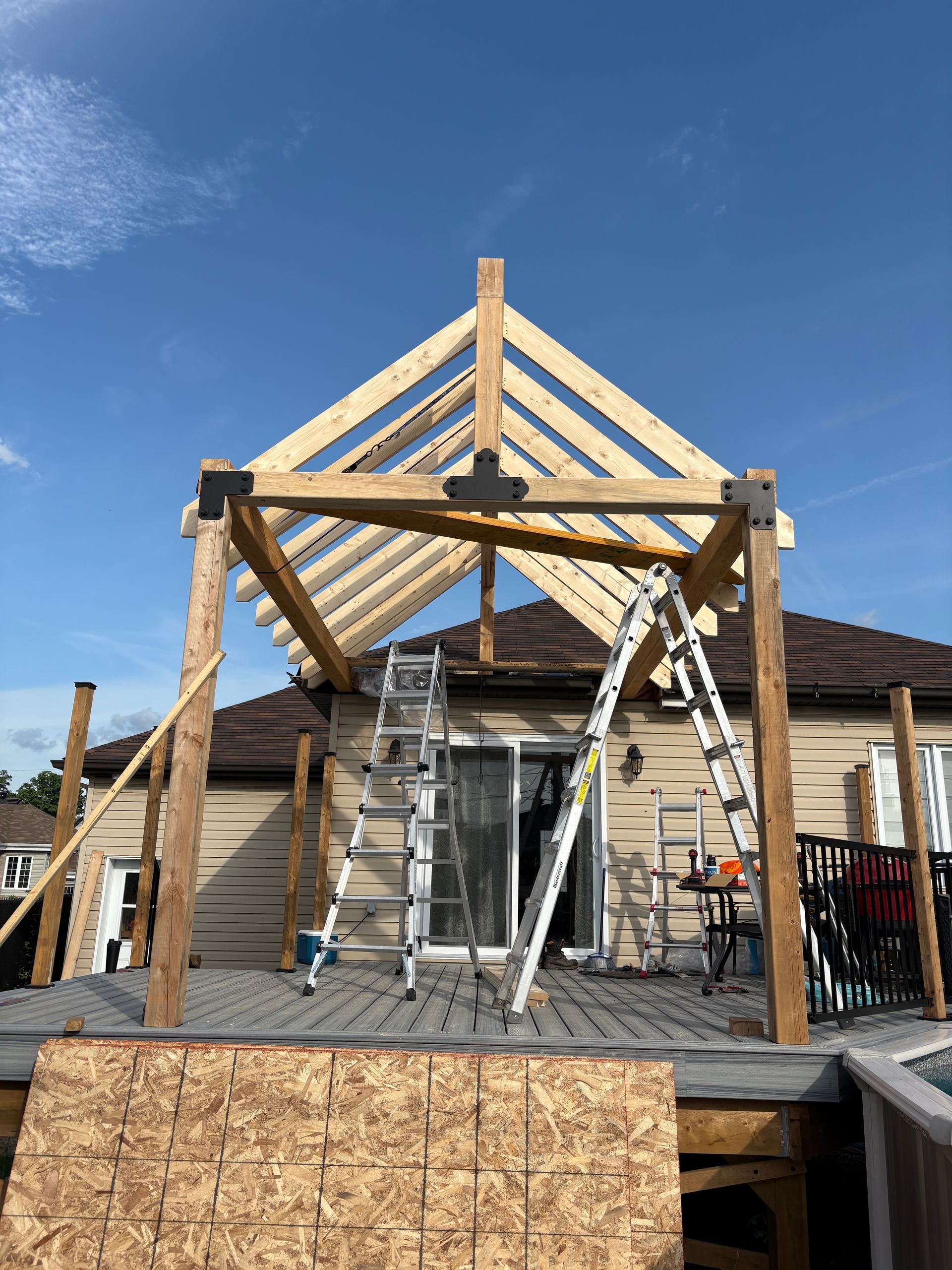 Construction d'une pergola en bois sur une terrasse, avec des échelles, devant une maison beige et un ciel bleu.