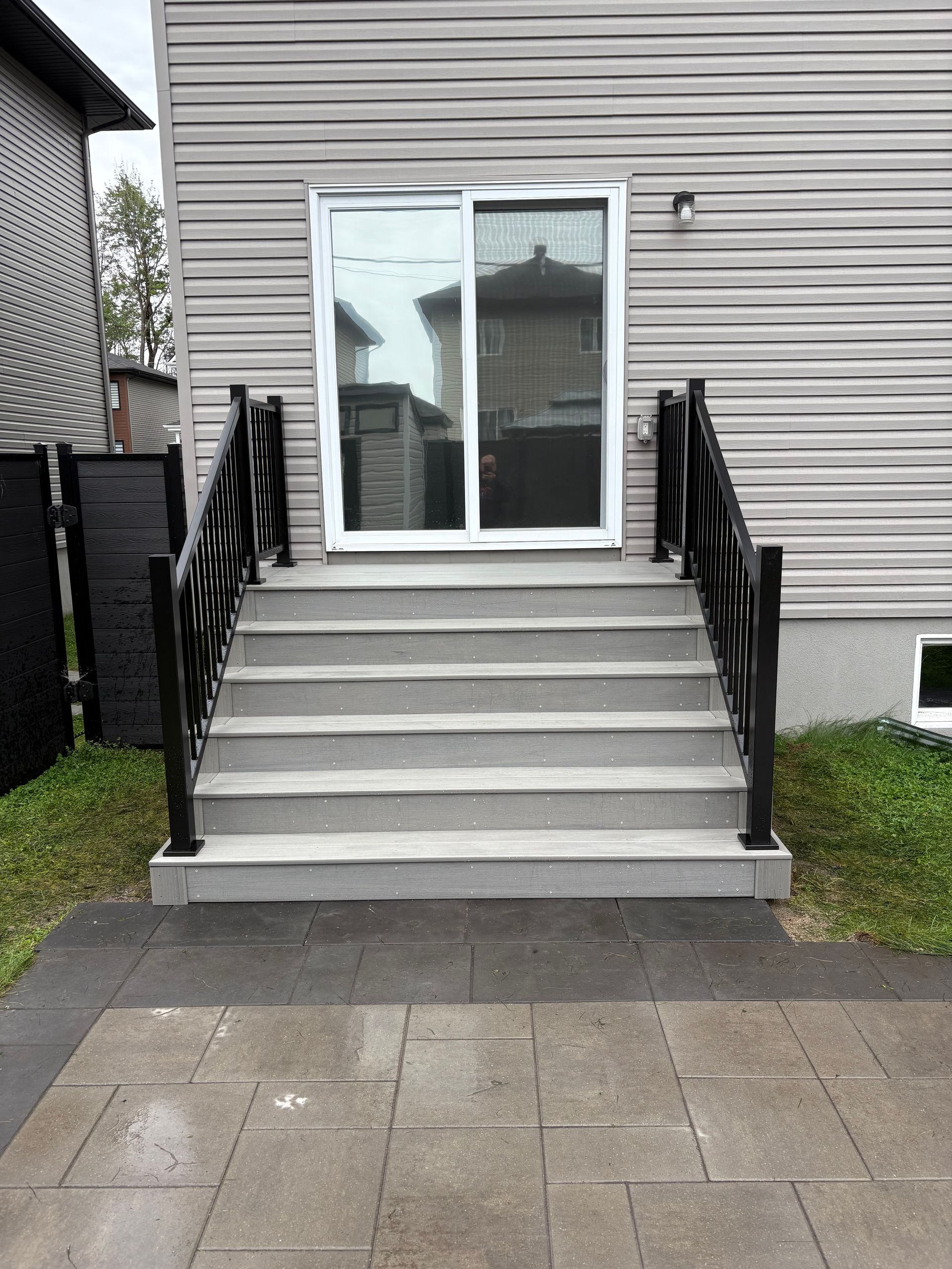 Des marches en béton gris mènent à une porte coulissante en verre. Des rambardes noires encadrent le tout.