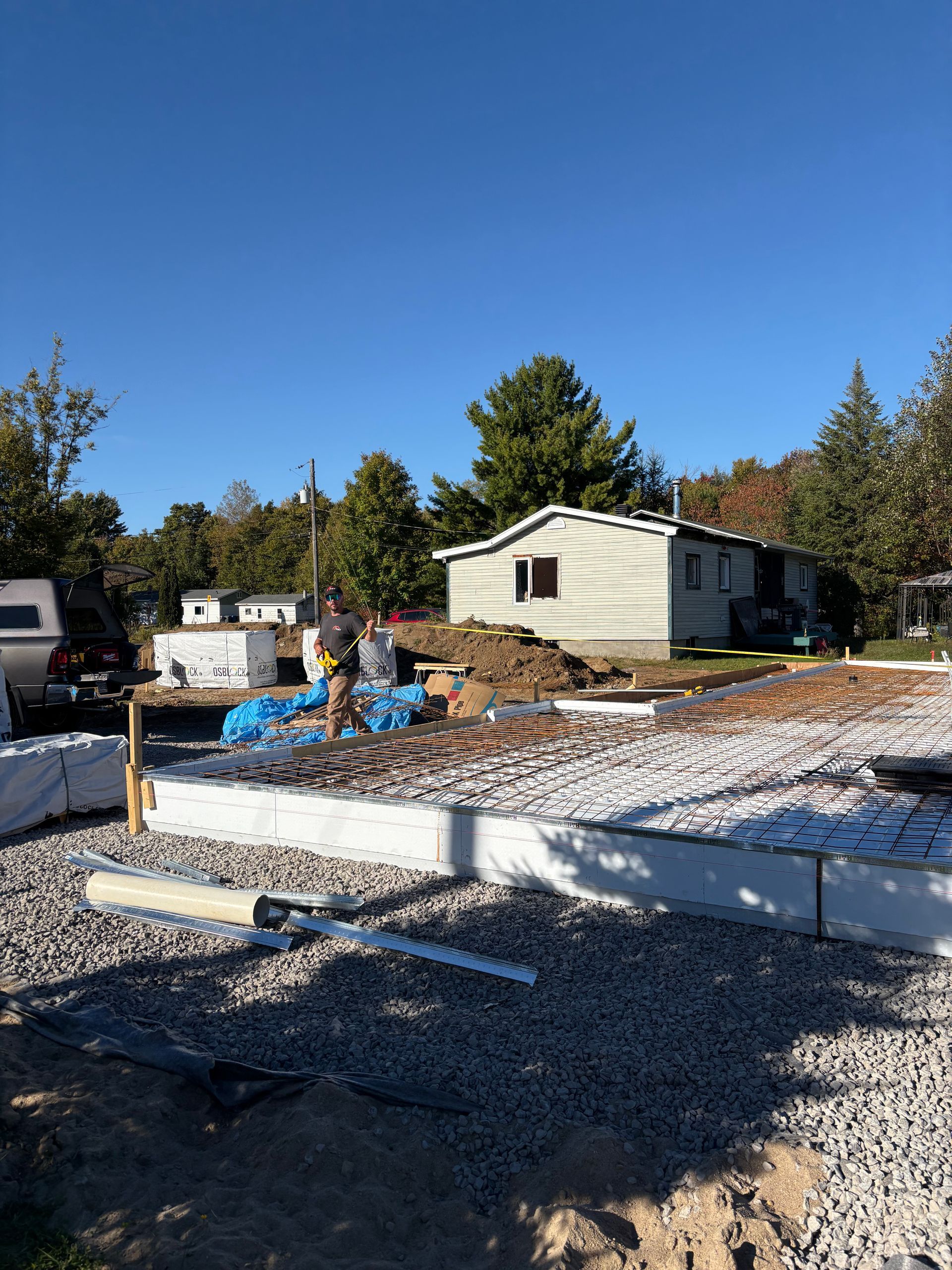 Chantier avec du gravier, des fondations et une petite maison en arrière-plan. Un ouvrier est visible.