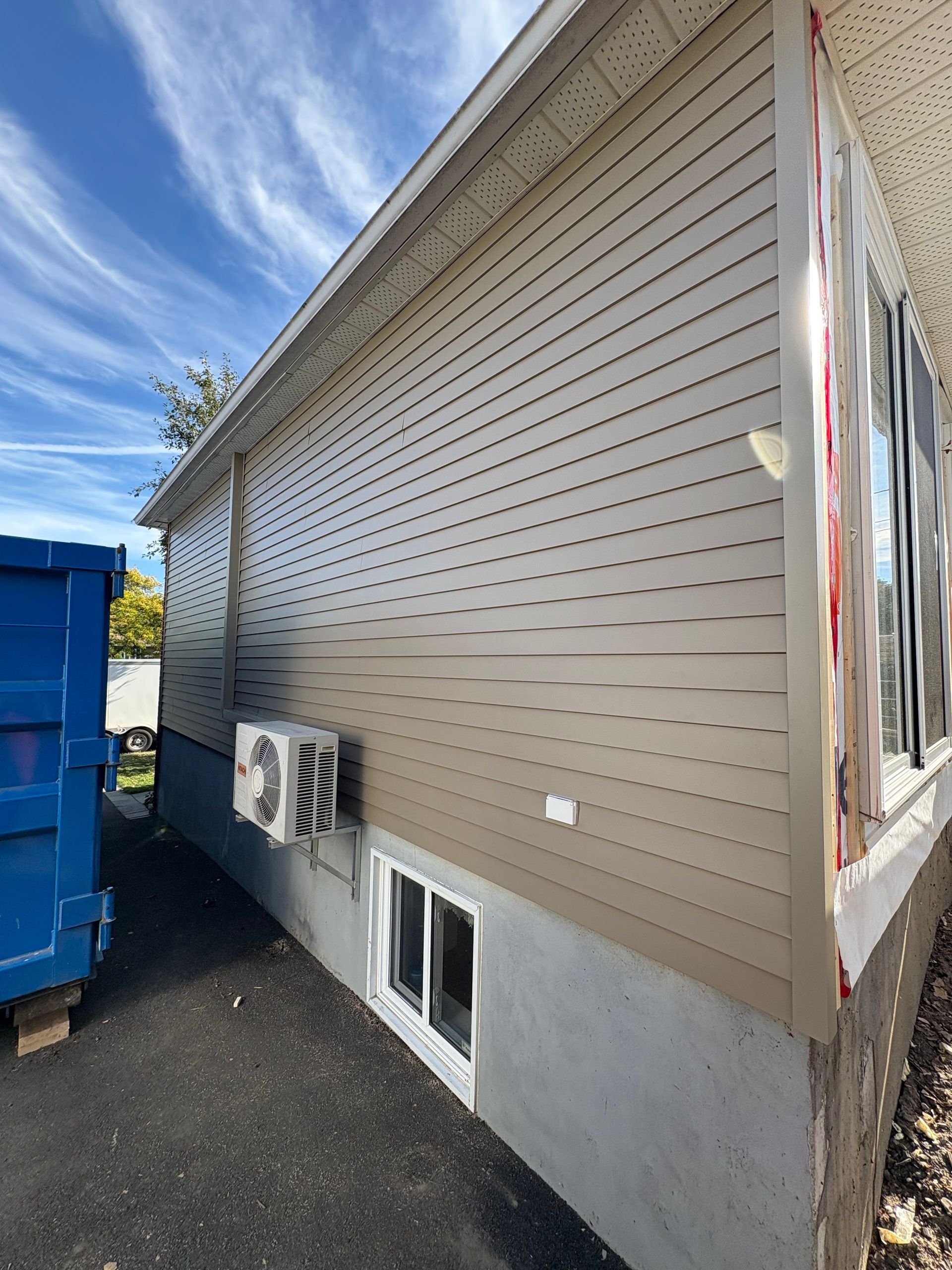 Façade d'une maison avec un revêtement beige et marron clair, une fenêtre et un climatiseur, à côté d'une benne à ordures bleue.