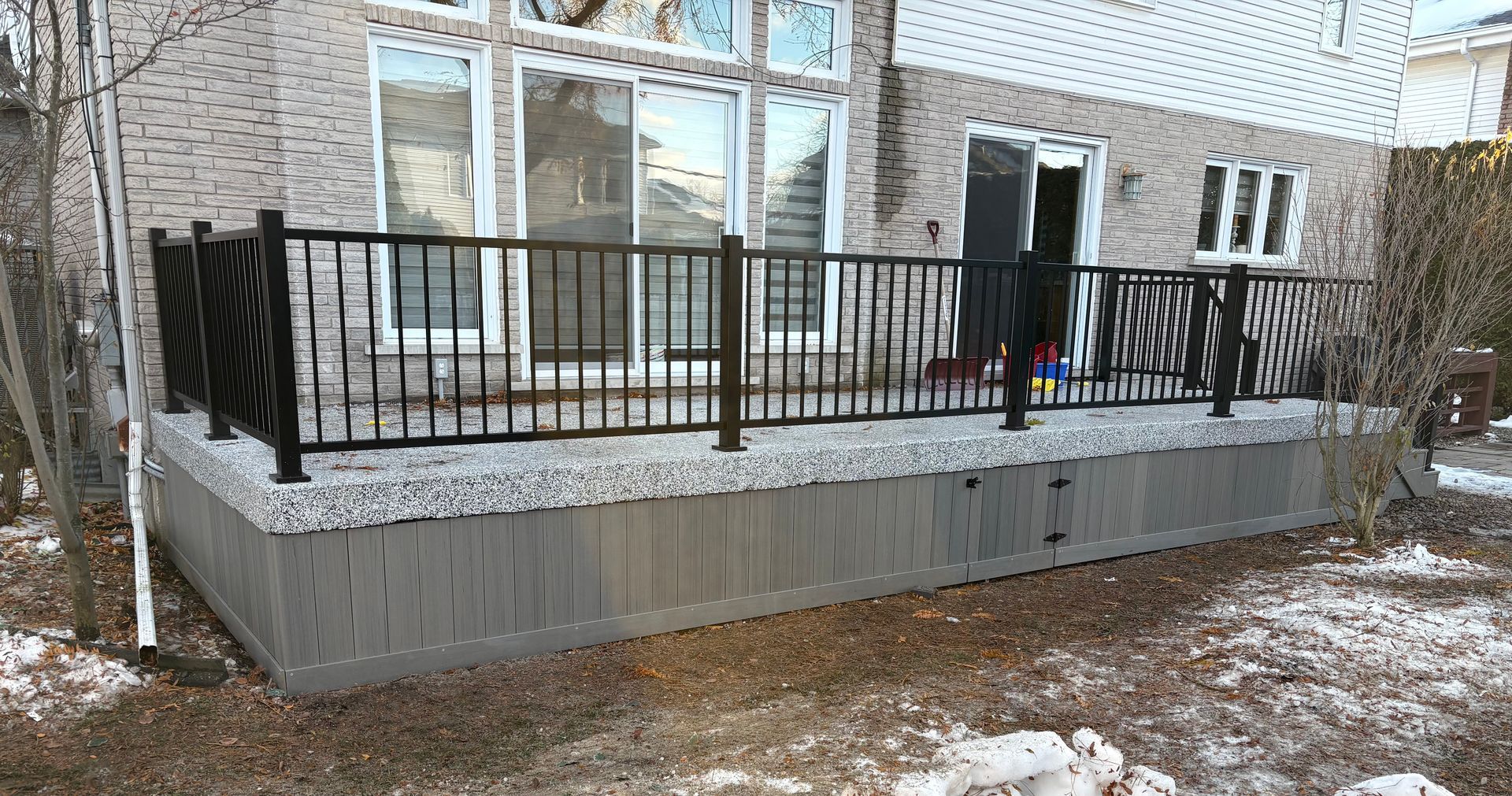 Terrasse avec balustrade noire et plinthes en composite gris, attenante à une maison en briques. Sol en gravier et neige.