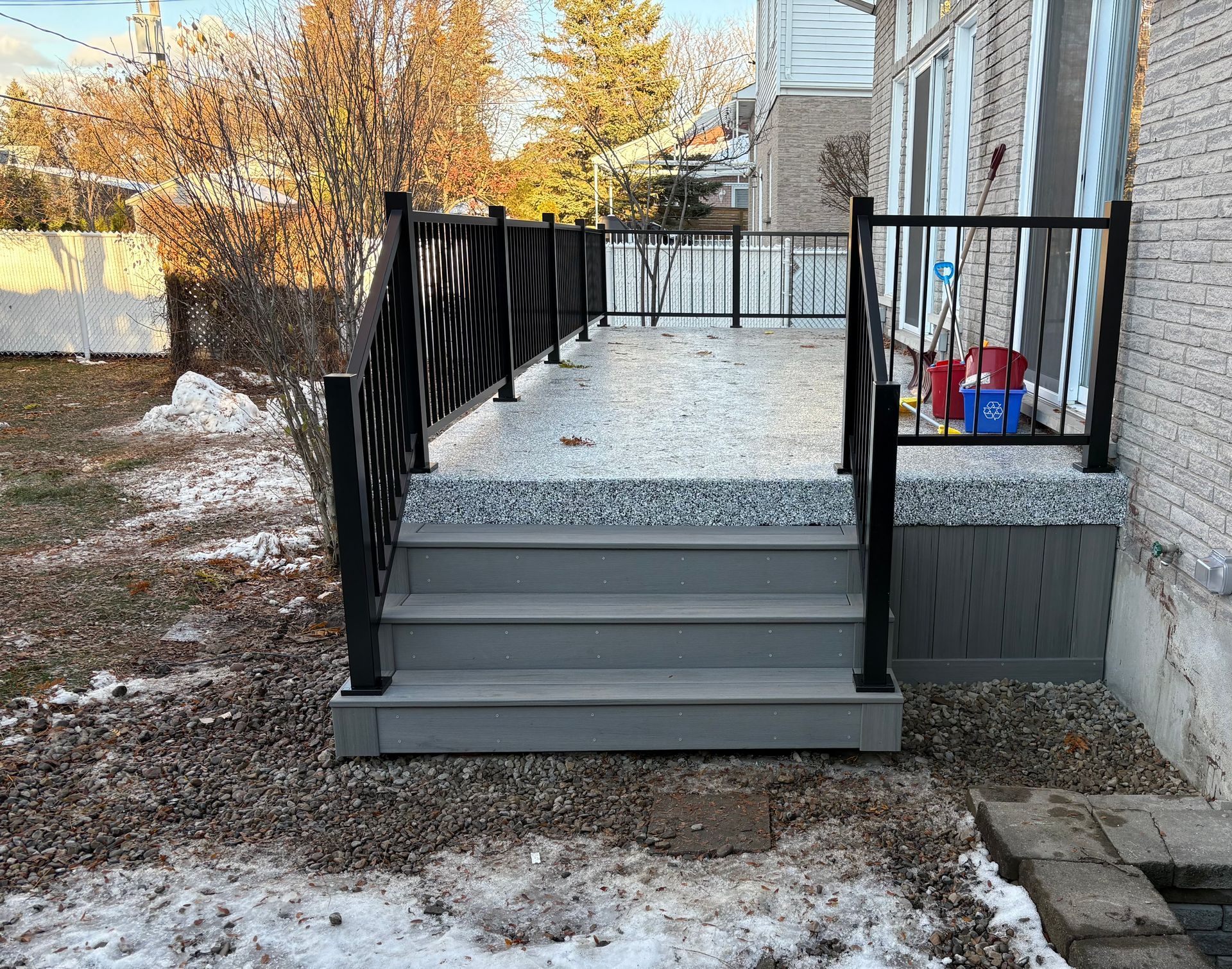 Terrasse en composite avec marches et rambarde noire. Paysage d'hiver : neige, sol gravillonné. Mur de briques à droite.