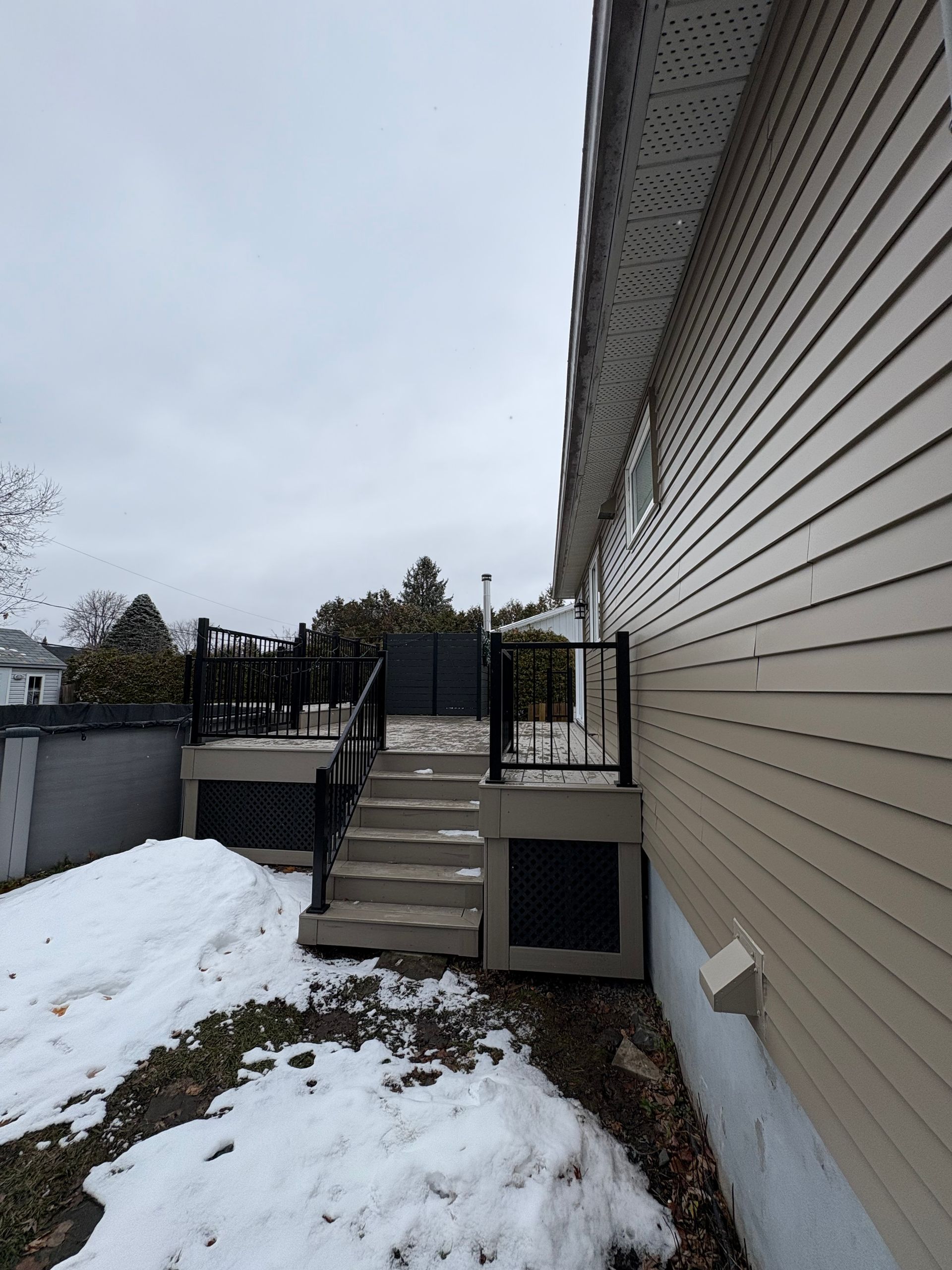 Une terrasse avec des marches et des rambardes noires jouxte une maison au bardage beige, la neige recouvre le sol.