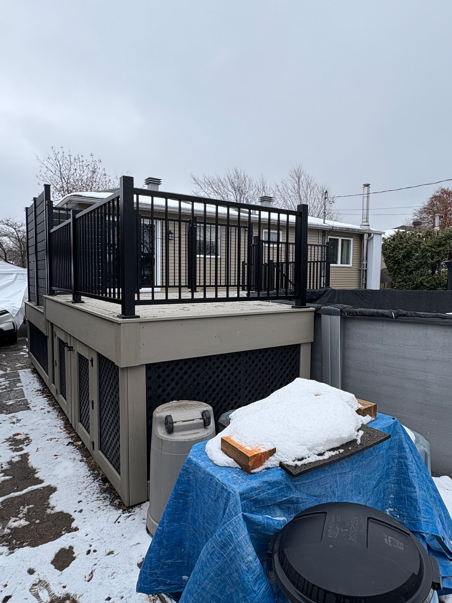 Terrasse avec rambarde noire sous la neige, donnant sur un jardin avec un objet recouvert d'une bâche et une piscine.