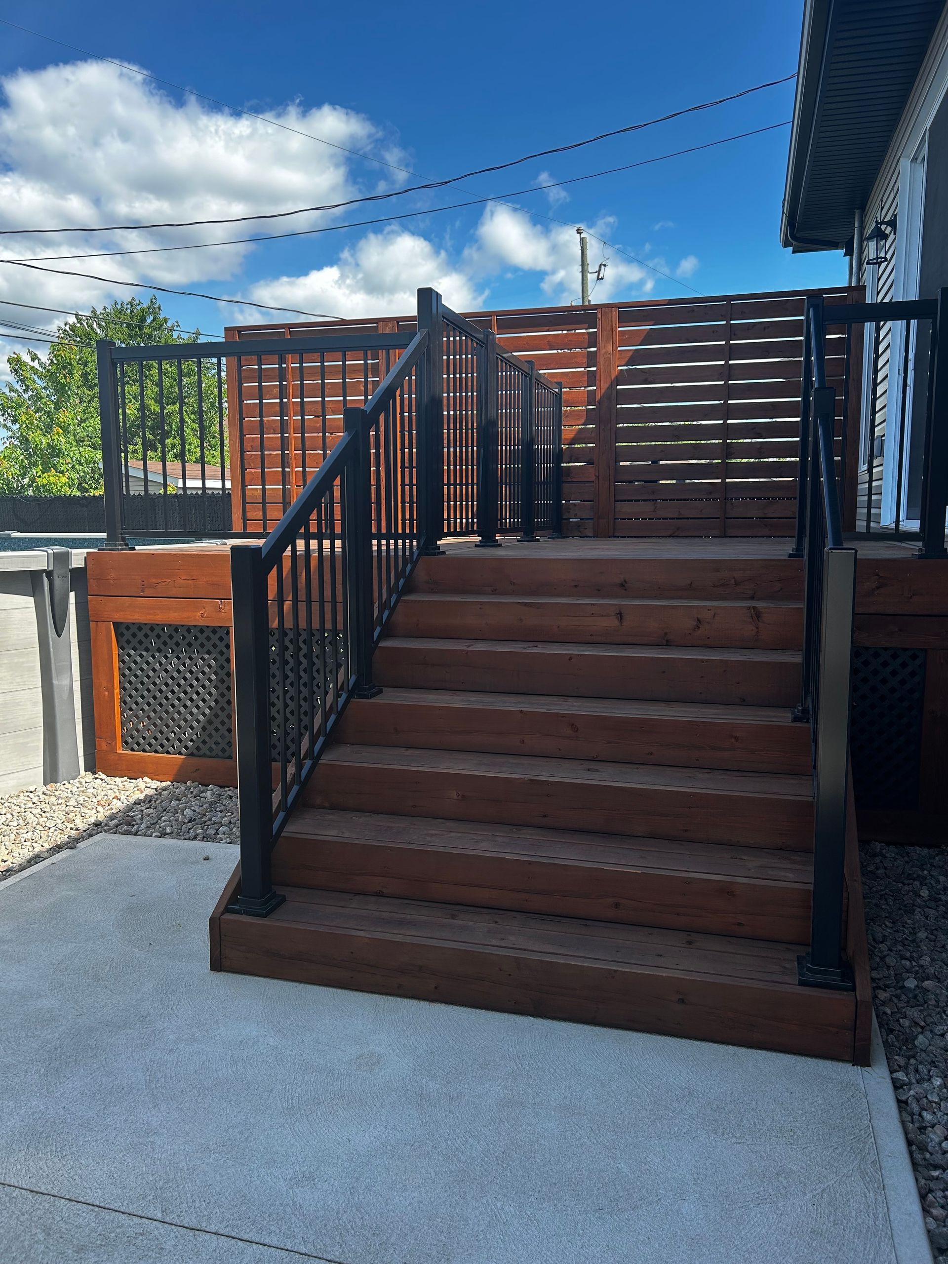 Terrasse en bois avec rambardes noires et marches menant à un paravent à claire-voie, sous un ciel bleu.