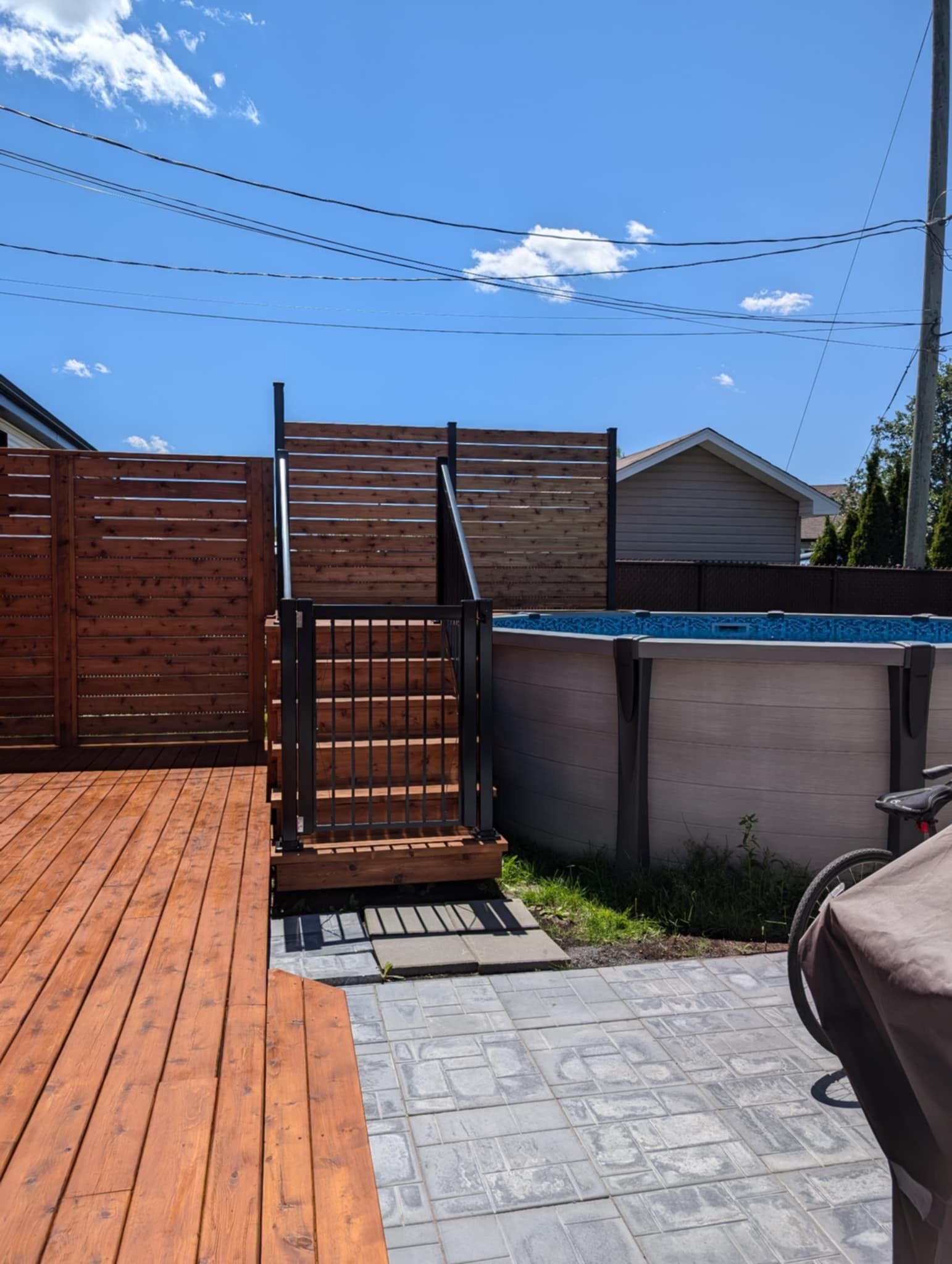 Terrasse en bois avec escalier menant à une piscine hors sol. Clôture en bois brun autour de la piscine. Ciel bleu au-dessus.
