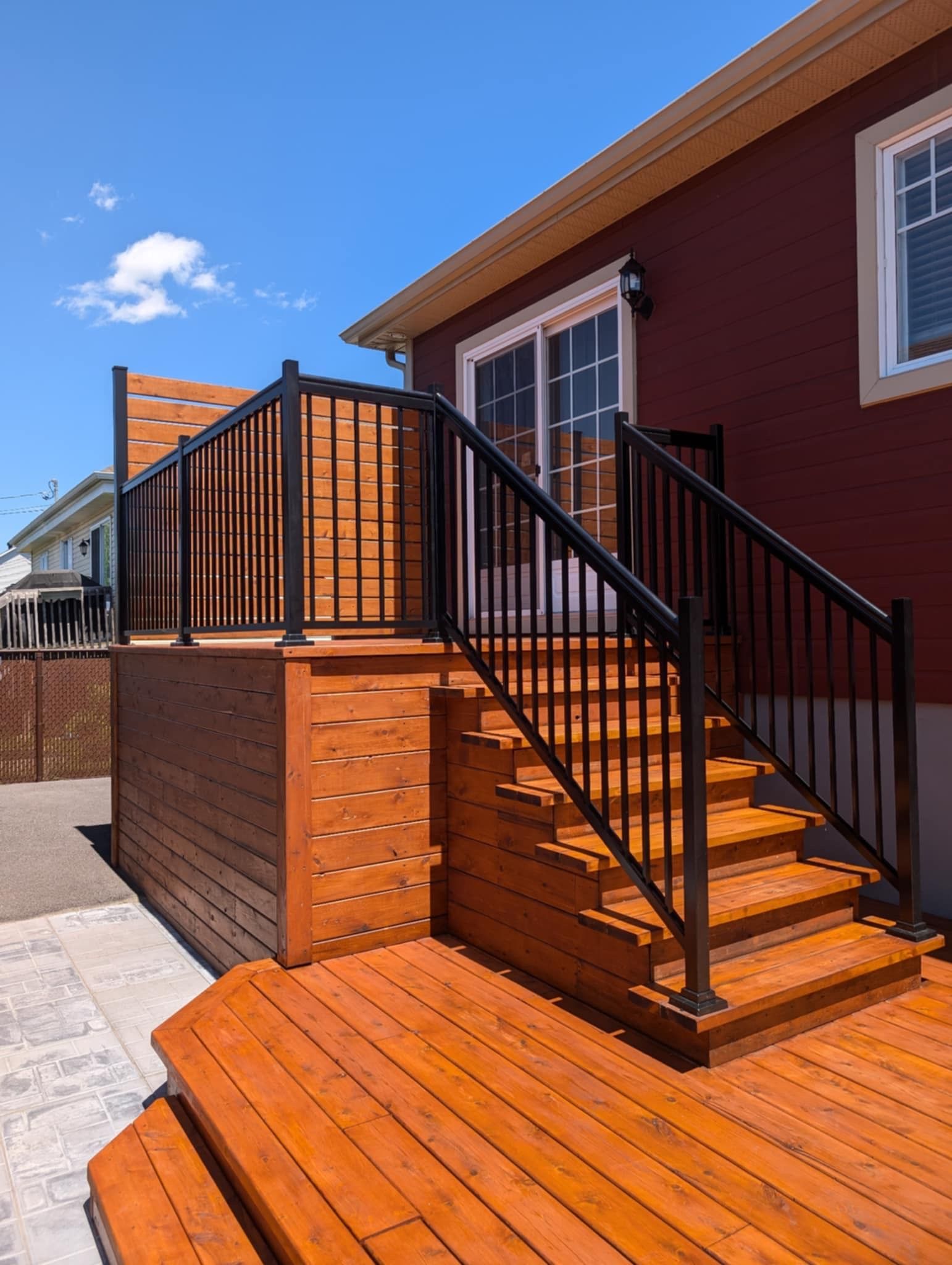 Terrasse en bois avec rambarde noire et escalier menant à une maison rouge avec une porte coulissante en verre.