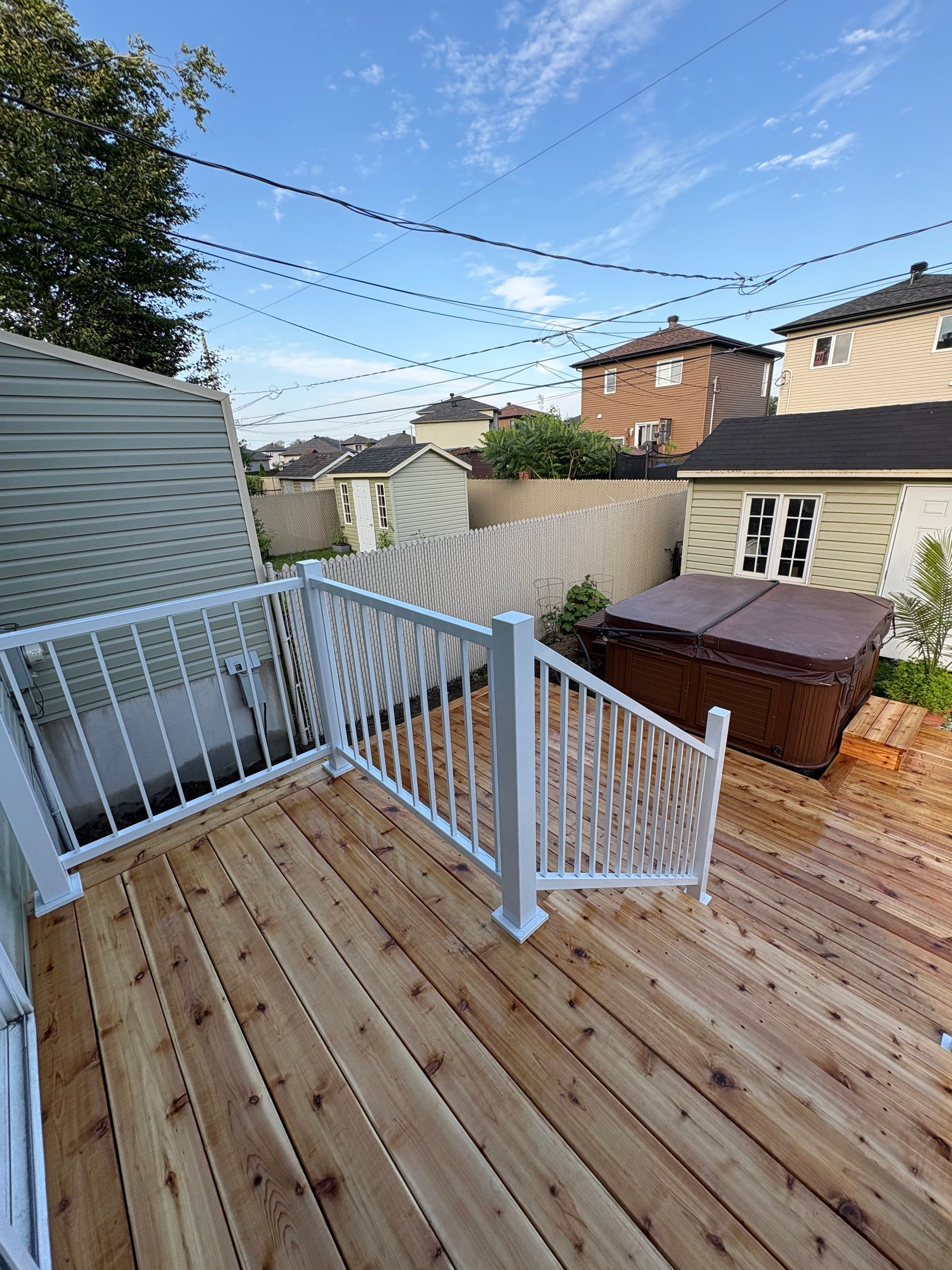 Terrasse en bois avec rambarde blanche à côté d'un jacuzzi. Ciel bleu avec des lignes électriques au-dessus.