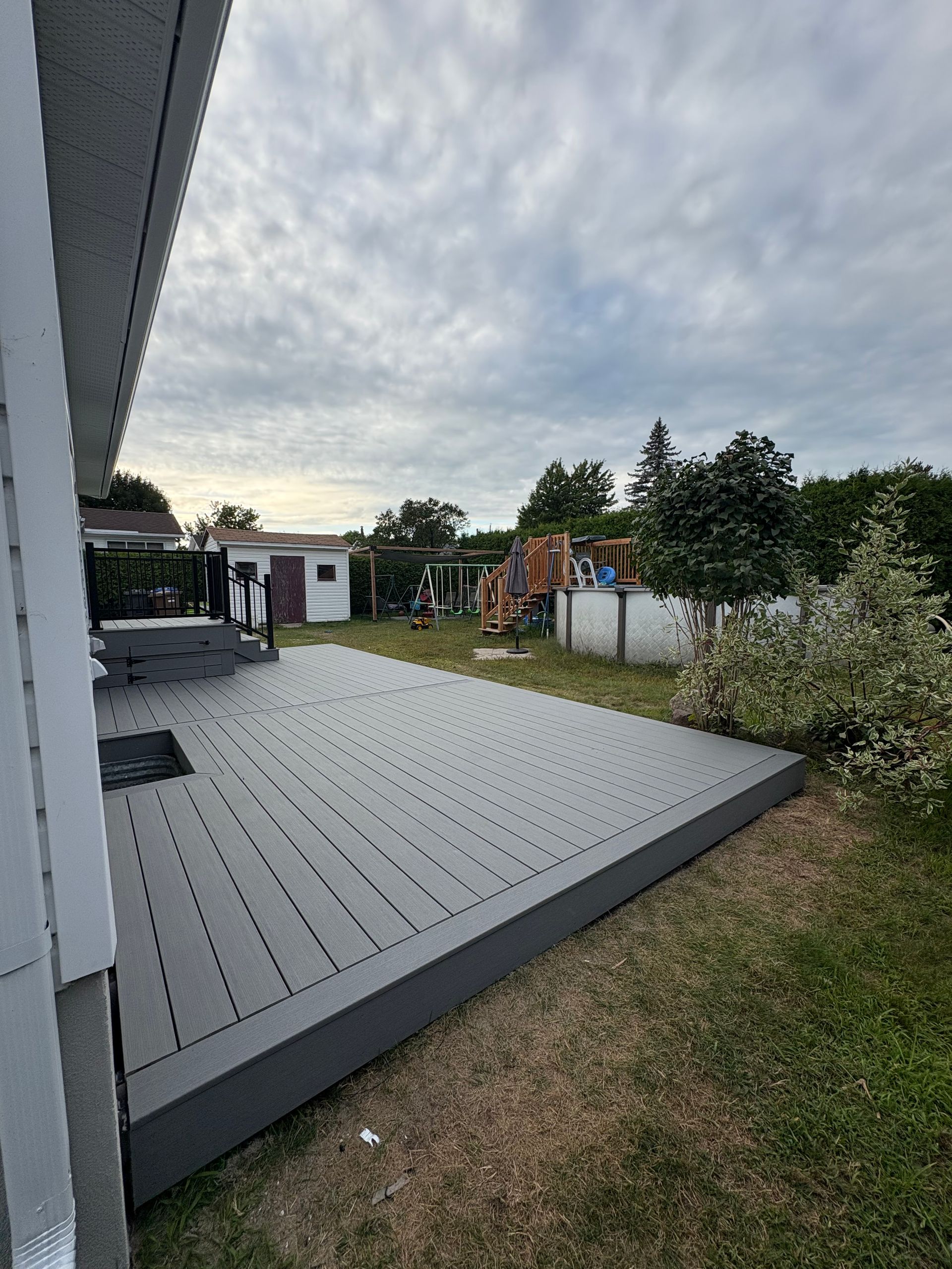 Une terrasse en composite gris s'ouvre sur un jardin gazonné, sous un ciel nuageux.