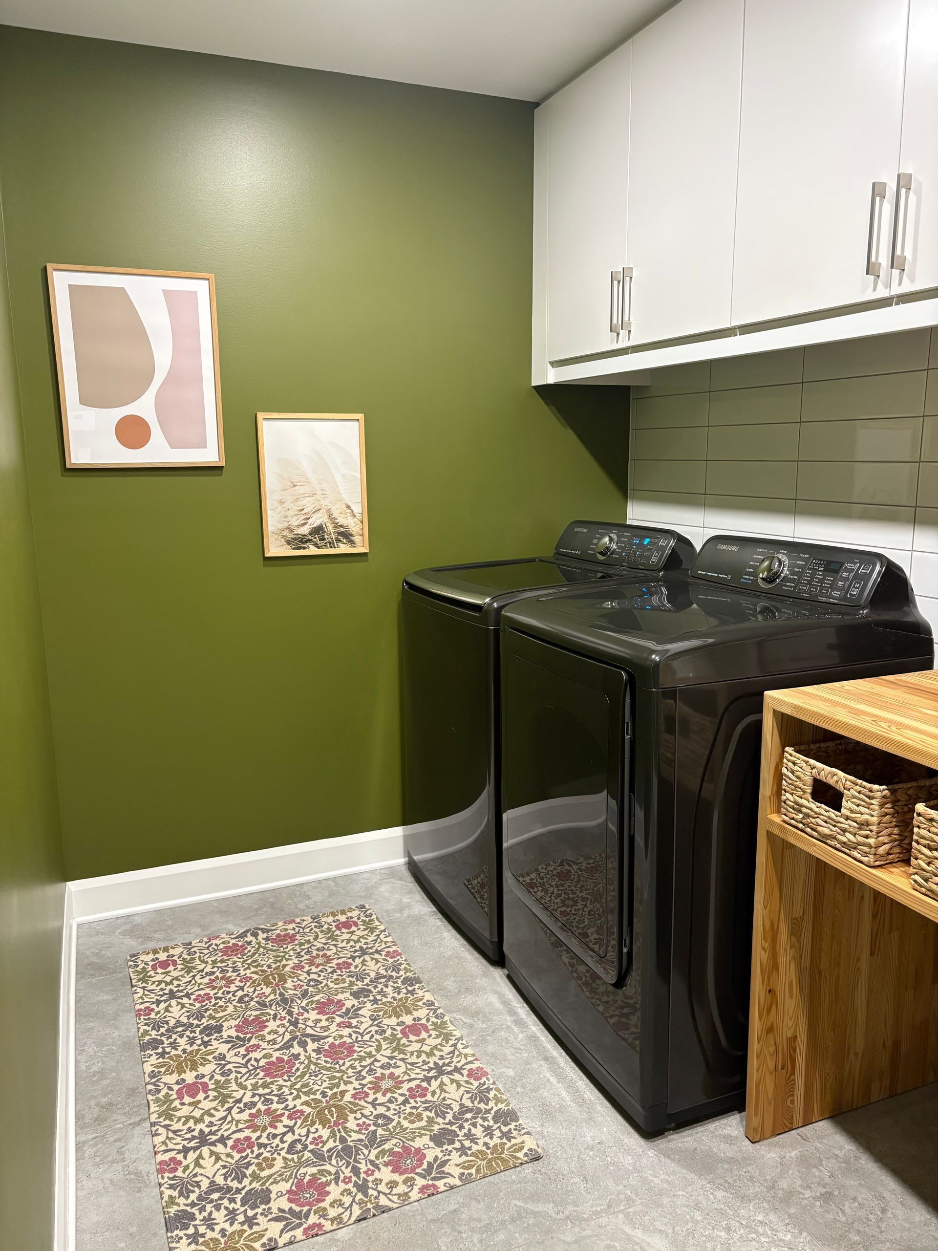 Buanderie avec un mur vert, lave-linge et sèche-linge noirs, et tapis à motifs floraux. Des placards blancs sont installés au-dessus.
