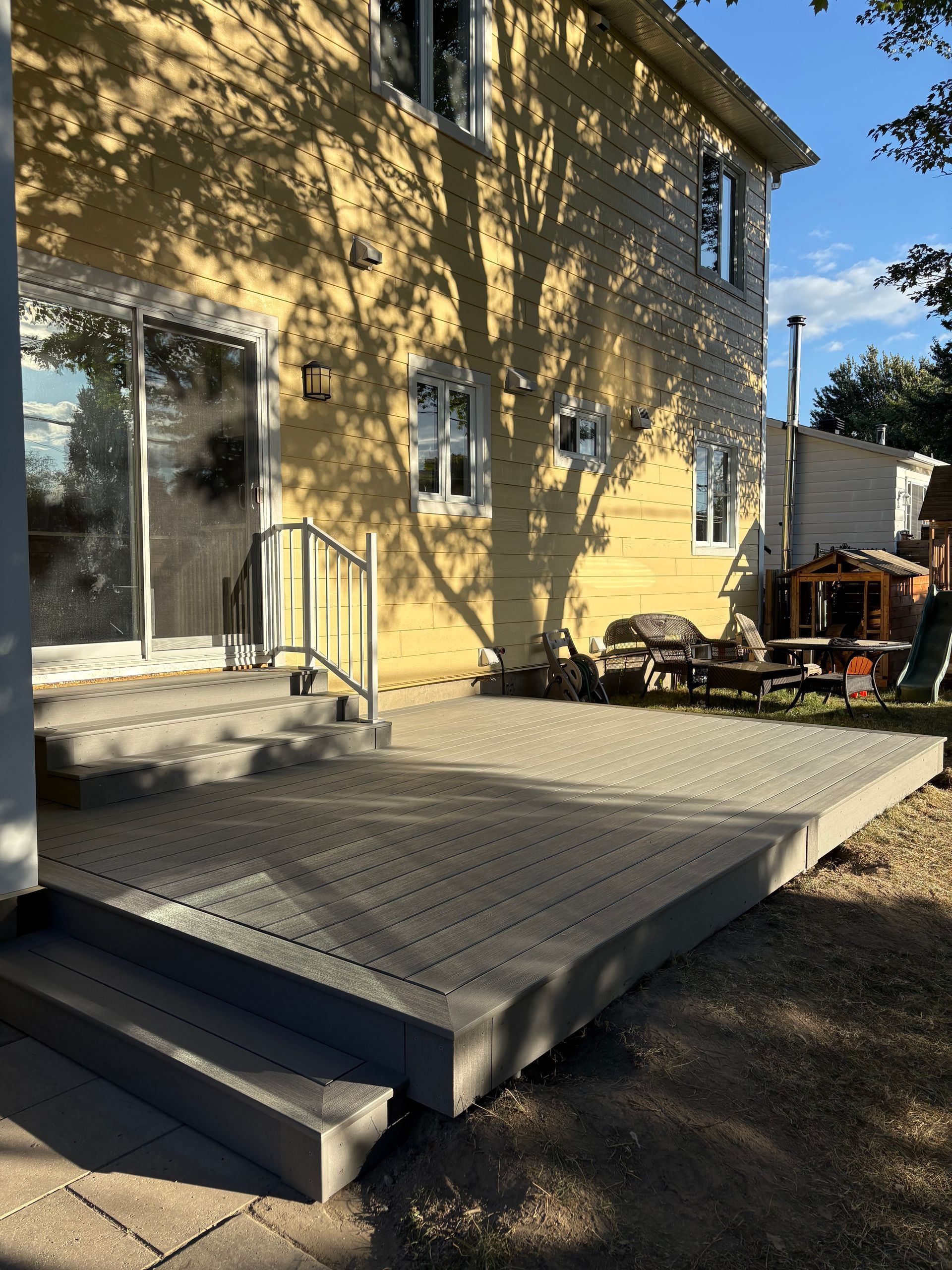 Terrasse en composite gris avec marches menant à une maison jaune à deux étages. Ombres d'arbres sur la maison et la terrasse.