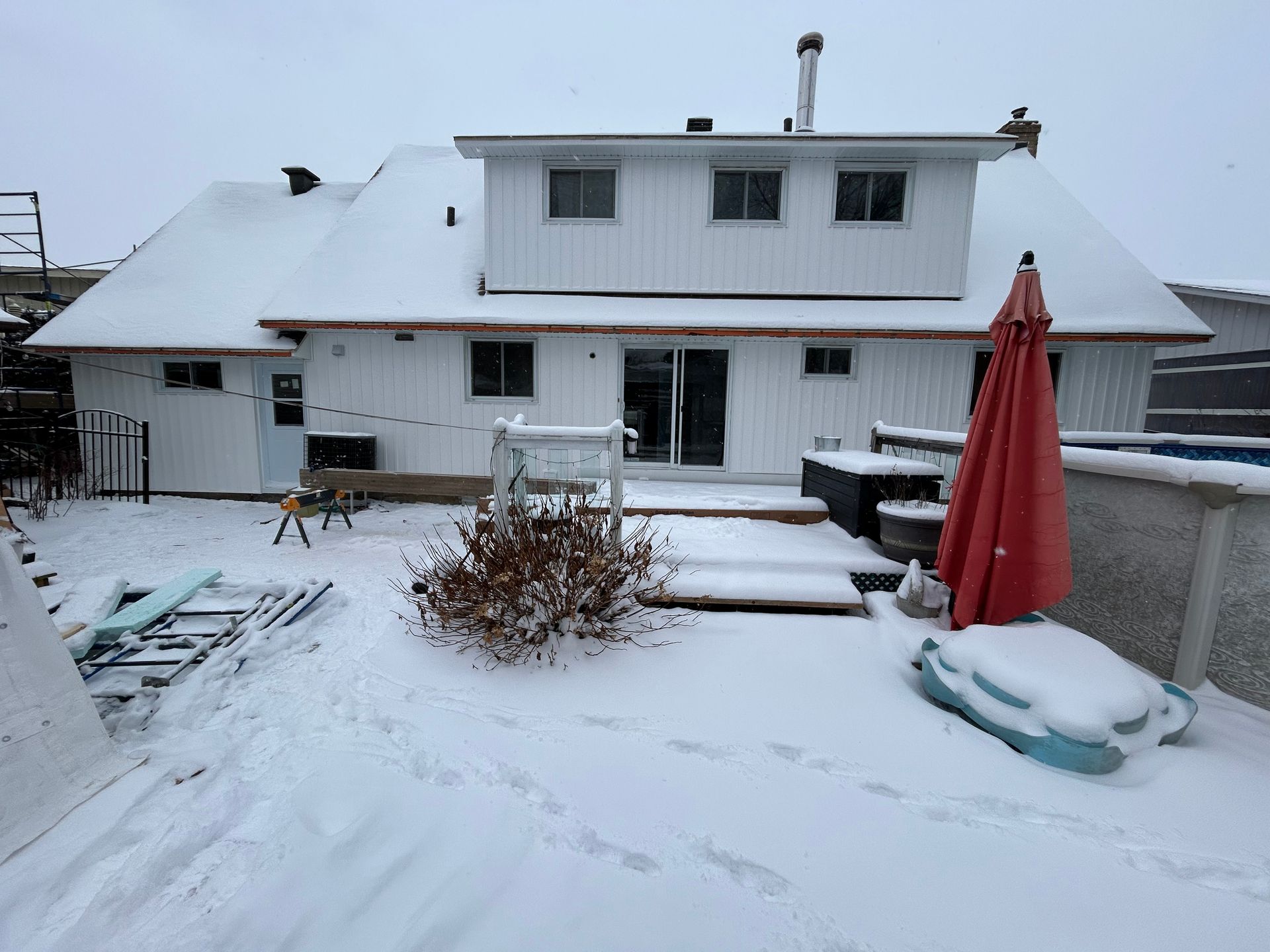 Jardin enneigé avec maison blanche, terrasse et parasol rouge. Scène hivernale avec des empreintes de pas dans la neige.