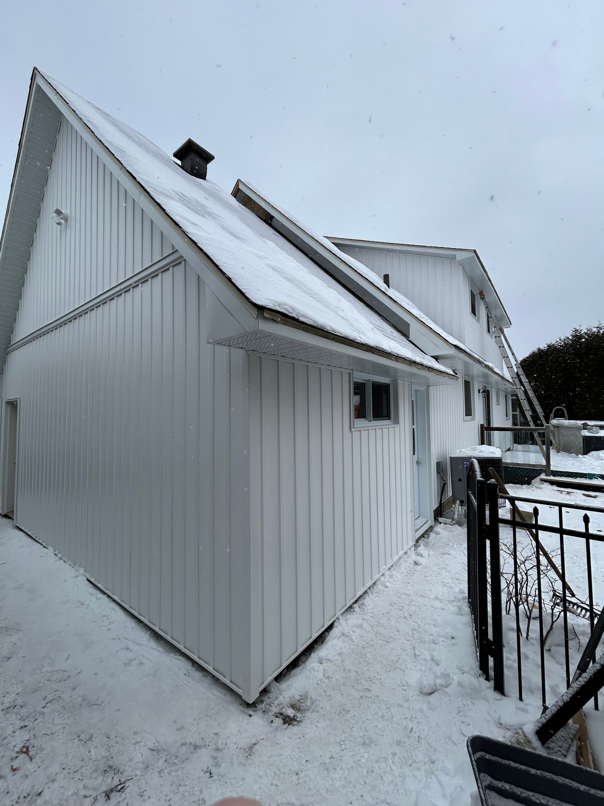 Maison blanche à l'extérieur, recouverte de neige. Toit à pignon avec cheminée noire, petite fenêtre et échelle.