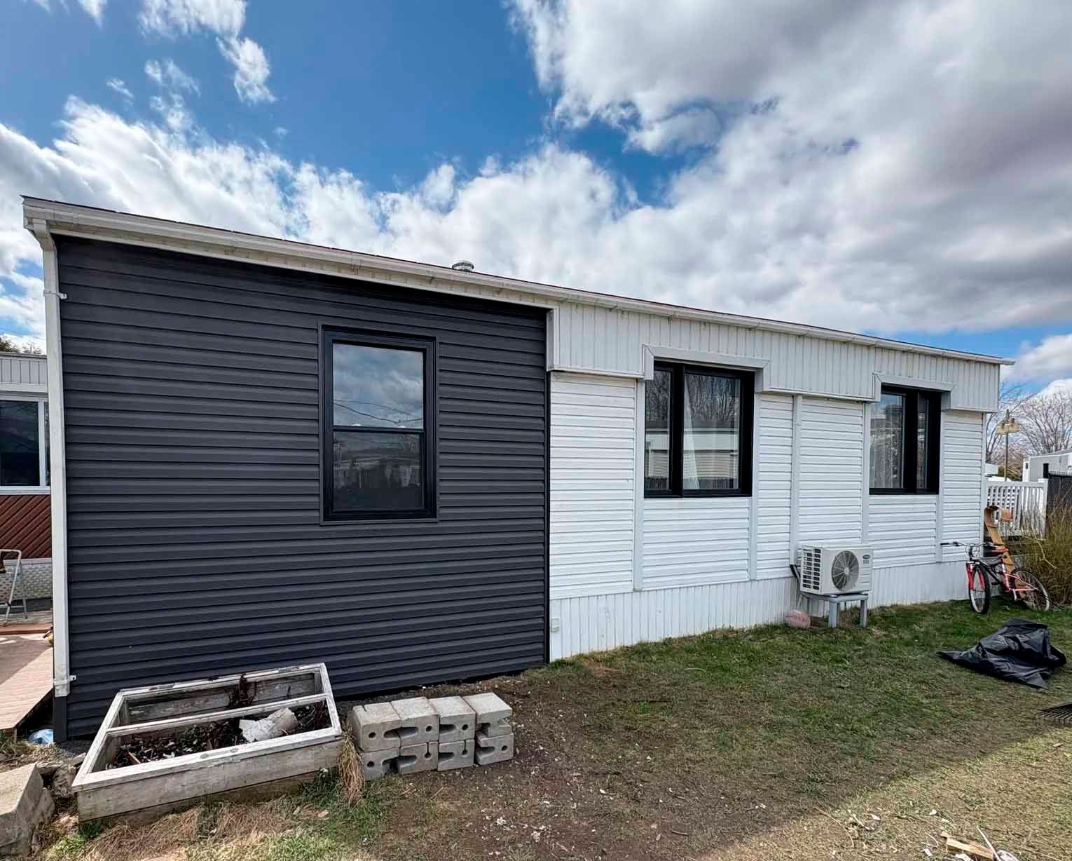 Extérieur d'une maison avec un revêtement noir d'un côté et blanc de l'autre, fenêtres et unité de climatisation.