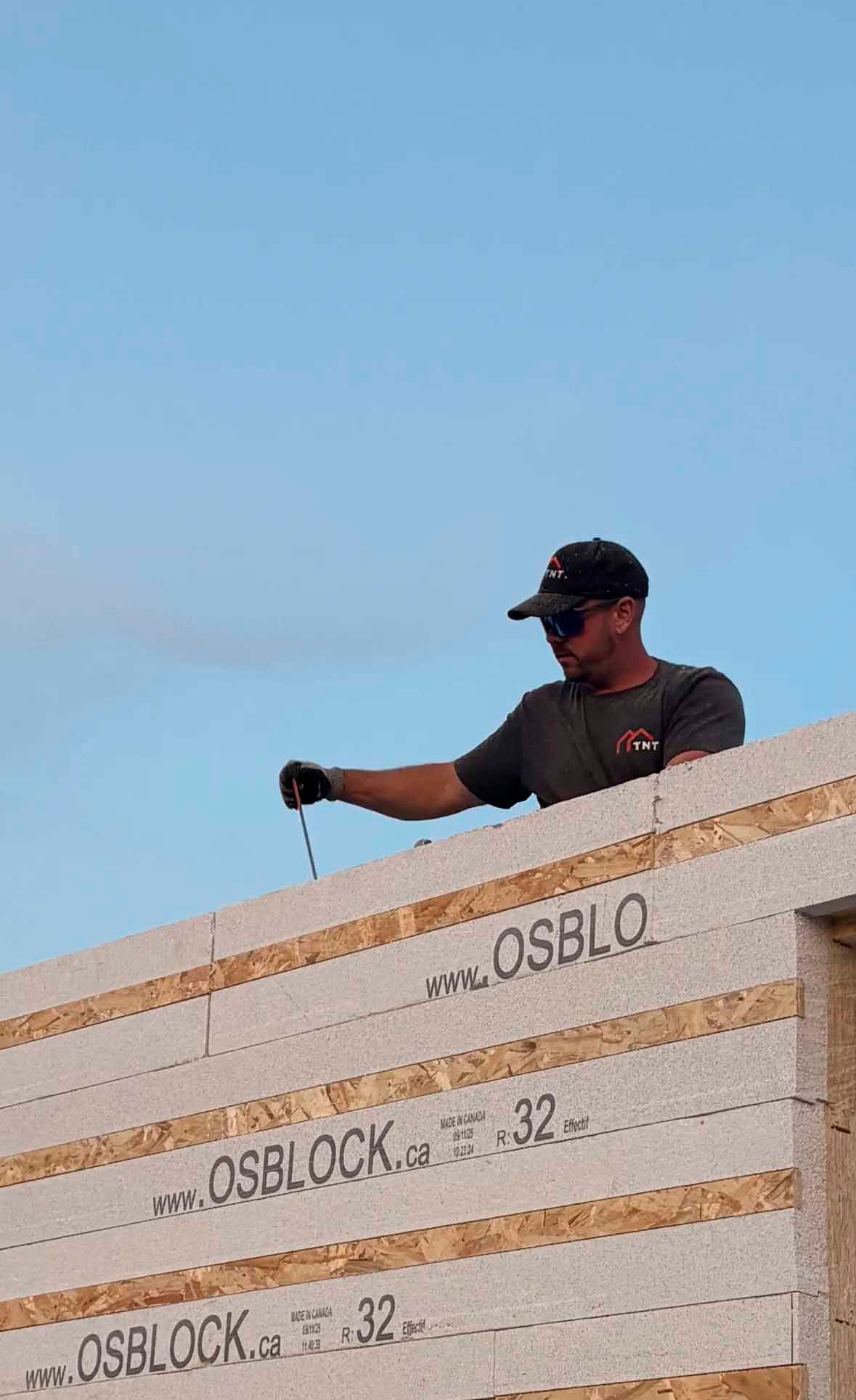 Ouvrier du bâtiment sur le toit, tenant un câble. Ciel bleu clair. Mur en blocs OSB visible.