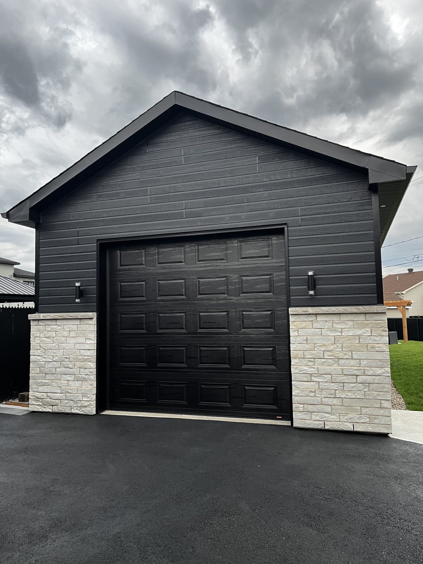 Garage noir avec des accents de pierre grise et allée asphaltée sur fond de ciel nuageux.