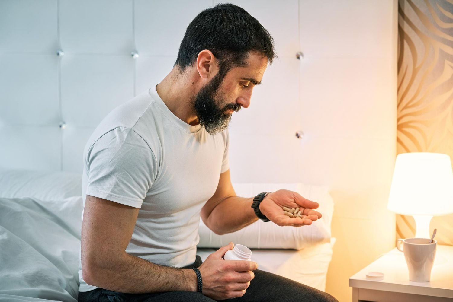 Man seated on a bed, holding pills from a bottle, bedside lamp glows.