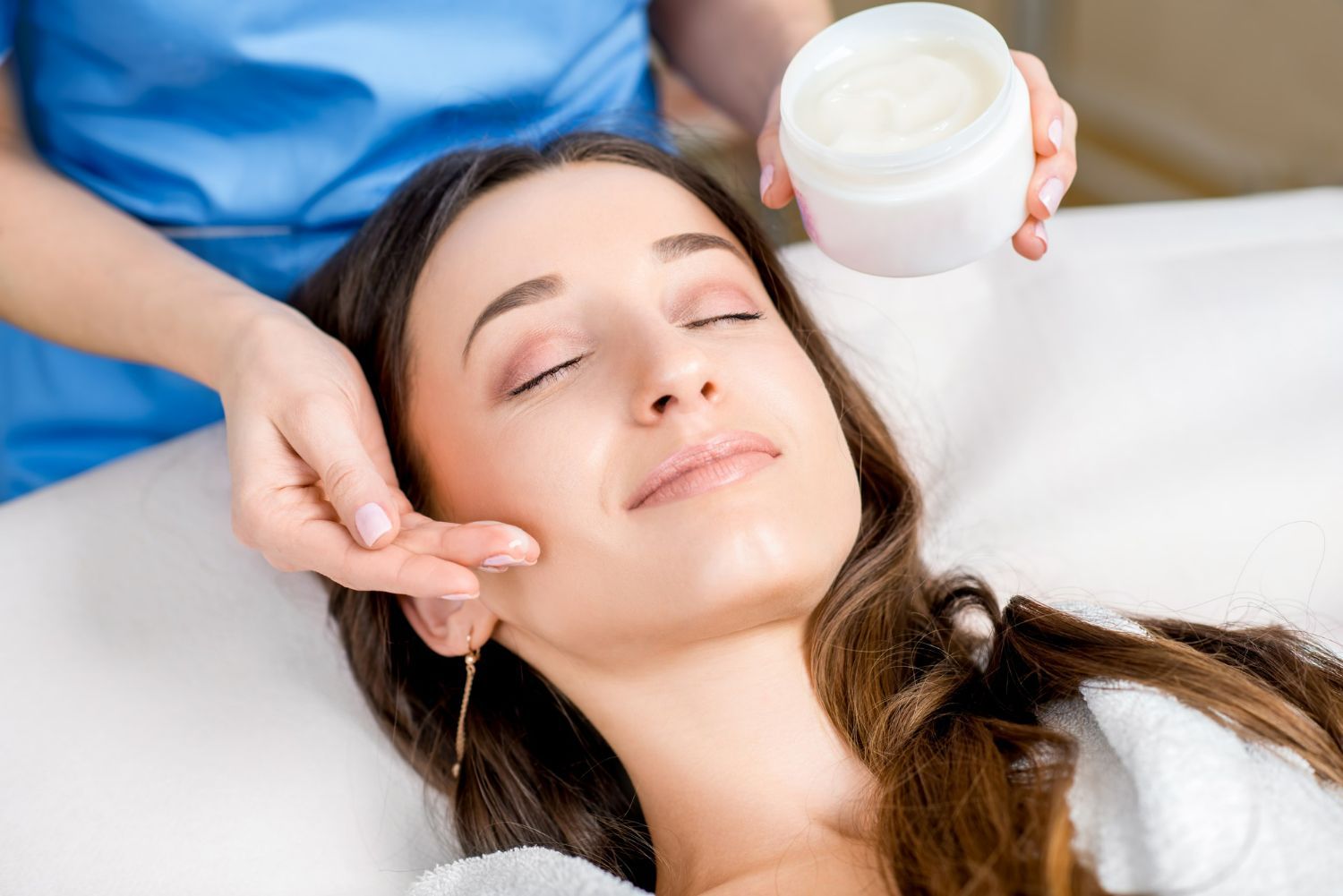 Woman receiving facial treatment; therapist holding cream jar and touching her cheek.