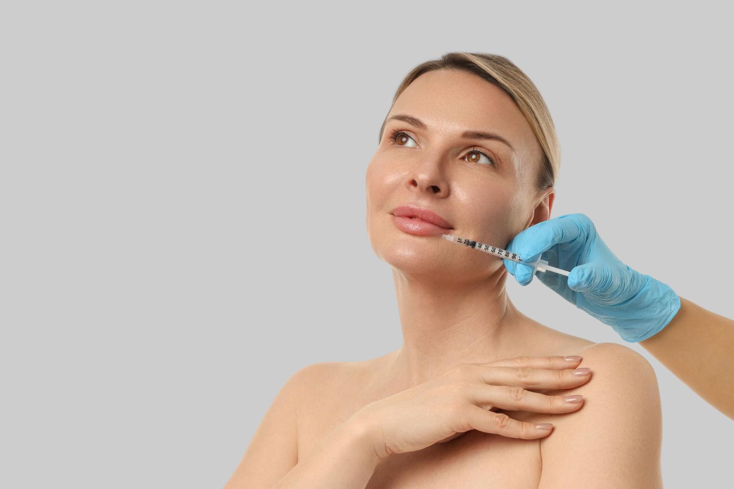 Woman receiving facial filler injection from a gloved practitioner against a gray background