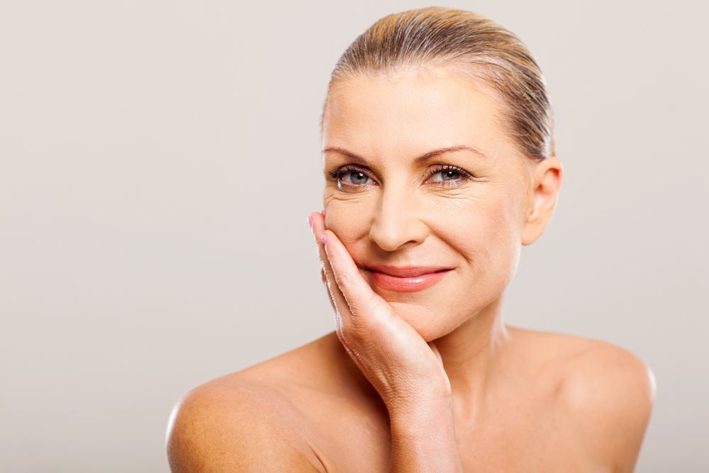 Woman with fair skin, smiling, hand on cheek, possibly having a facial treatment.