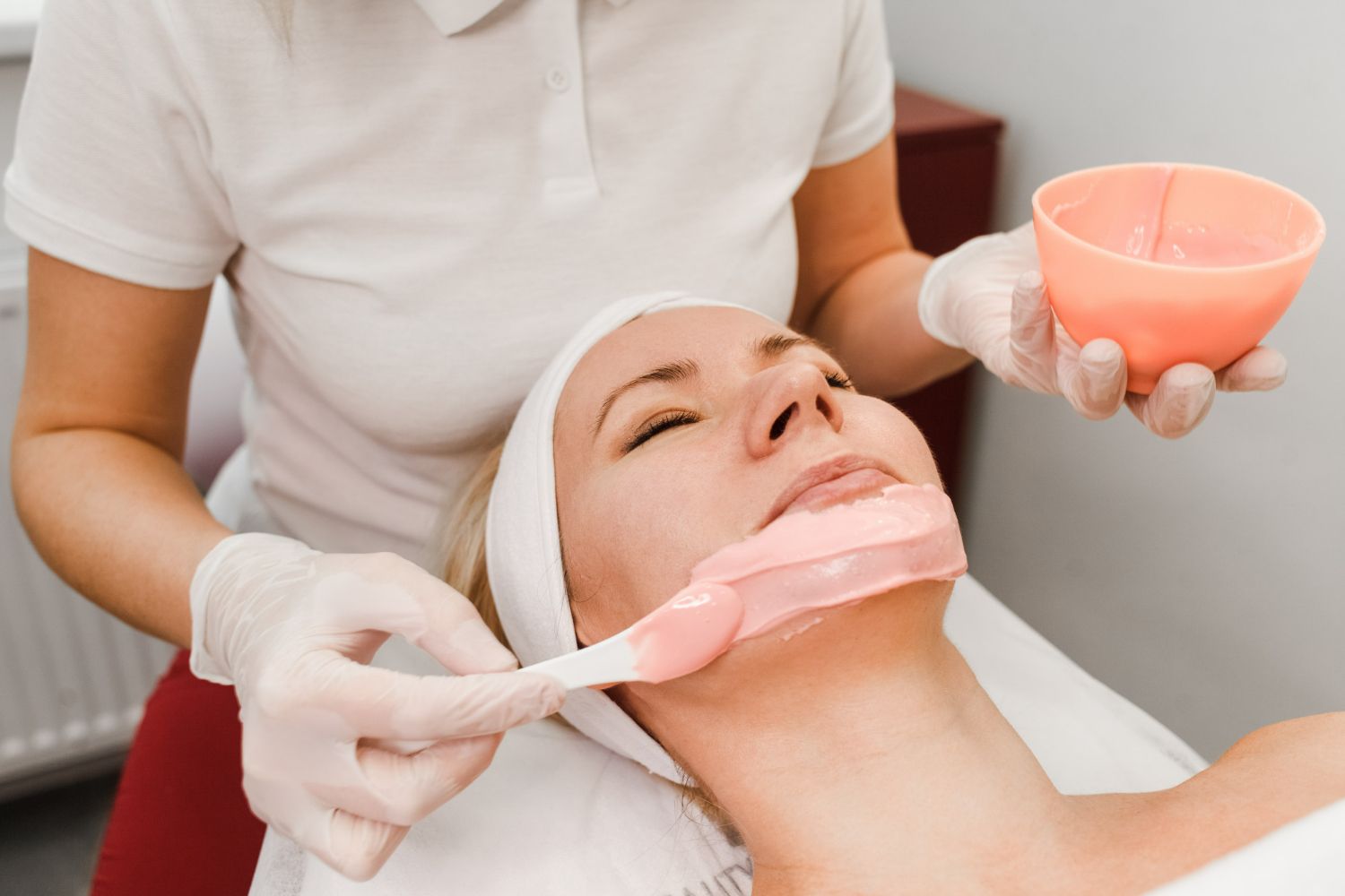 Woman receiving a facial treatment; pink mask applied to lower face.