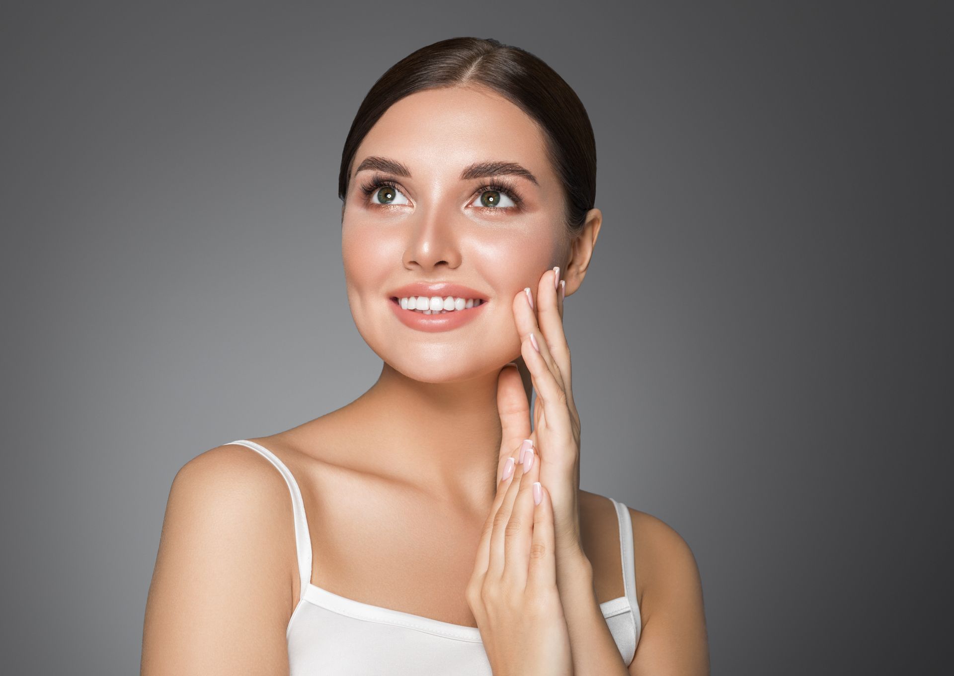 Woman with fair skin smiles, touching her cheek with her hands, looking upward against a grey background.