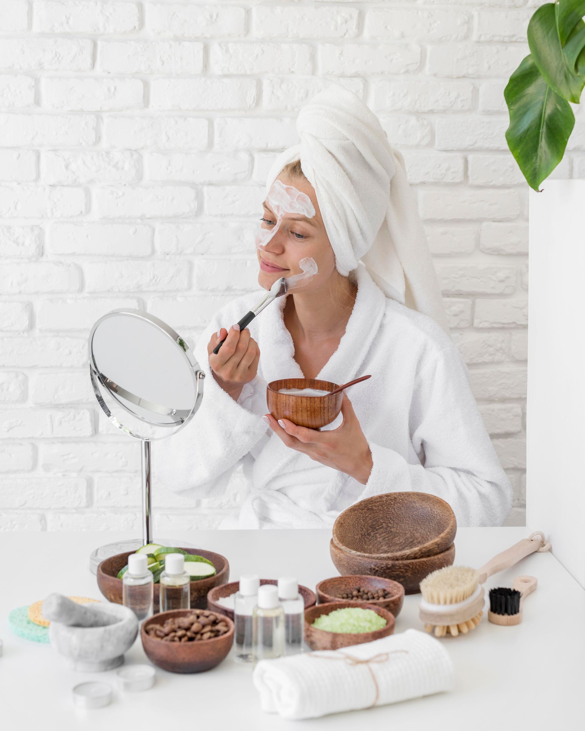 A woman with a towel wrapped around her head is applying a mask to her face.